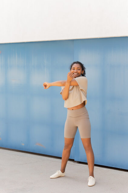 A woman in beige athletic wear stands on a sidewalk, stretching her arm across her chest in front of a blue wall.