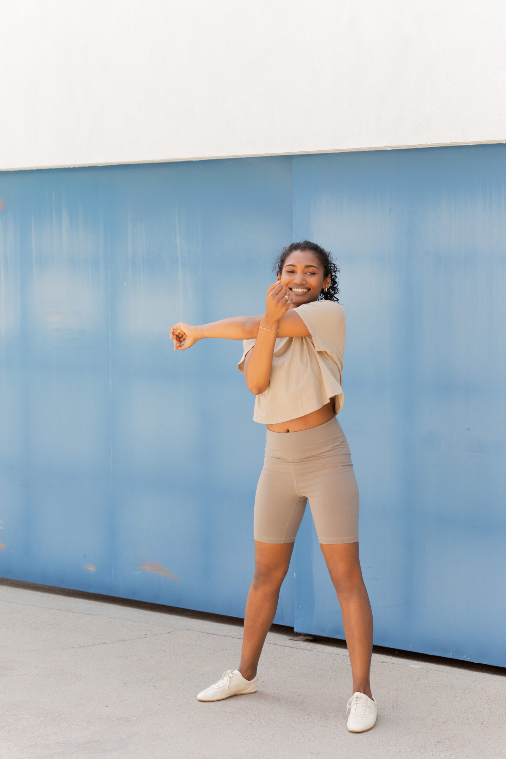 A woman in beige athletic wear stands on a sidewalk, stretching her arm across her chest in front of a blue wall.