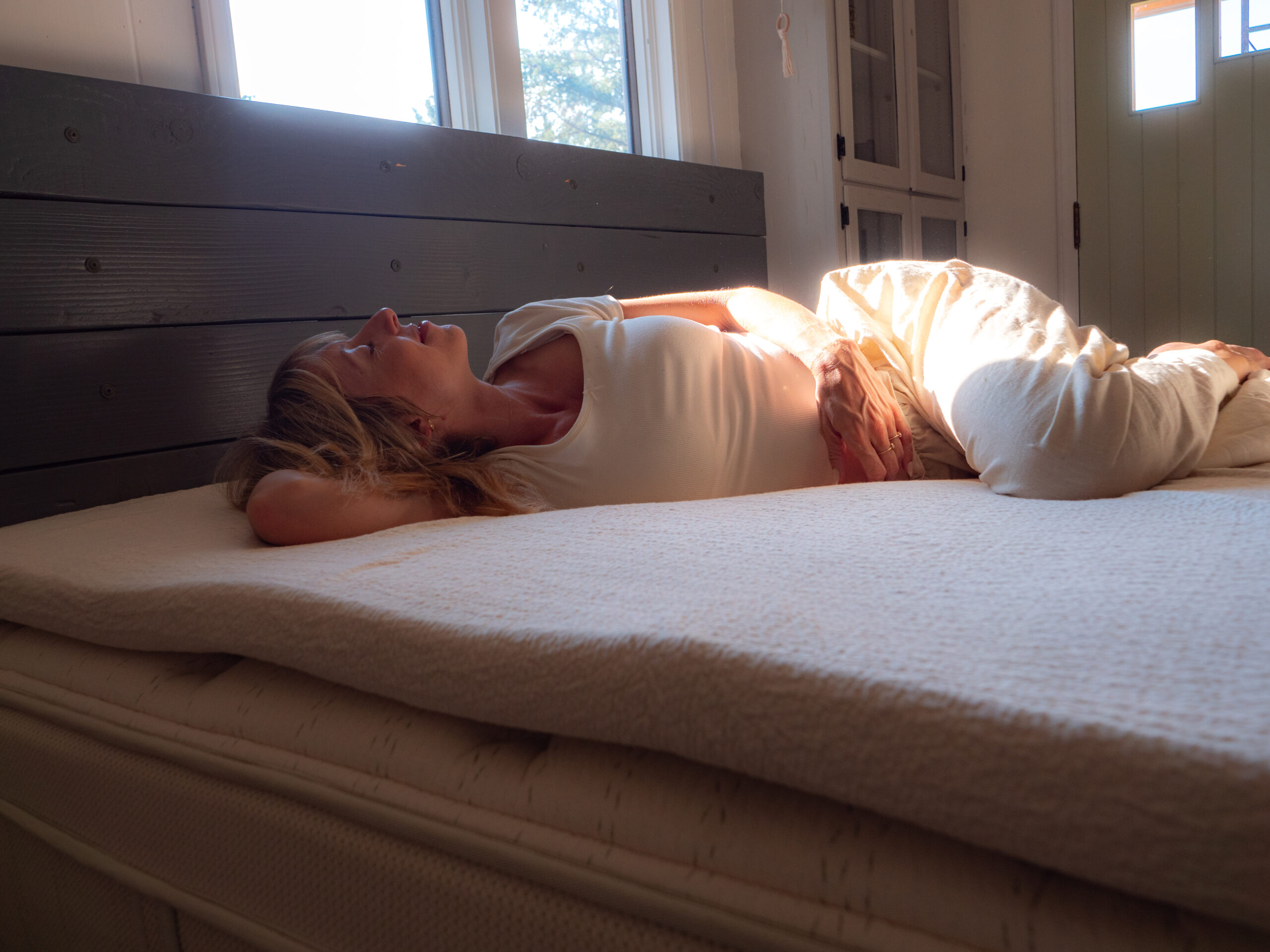 A woman in light clothing lies on her side on a bed in a sunlit room, with her hand resting on her abdomen and eyes closed.