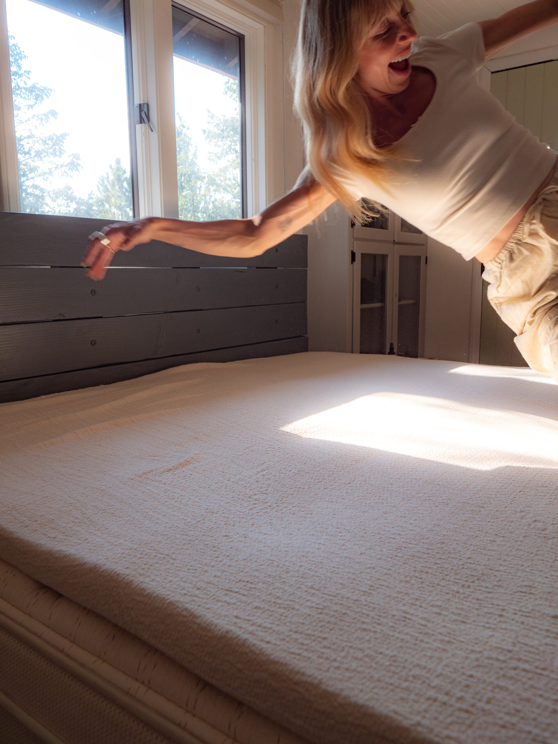 A woman in a white shirt and beige pants is falling or jumping onto a bed in a sunlit bedroom with large windows.