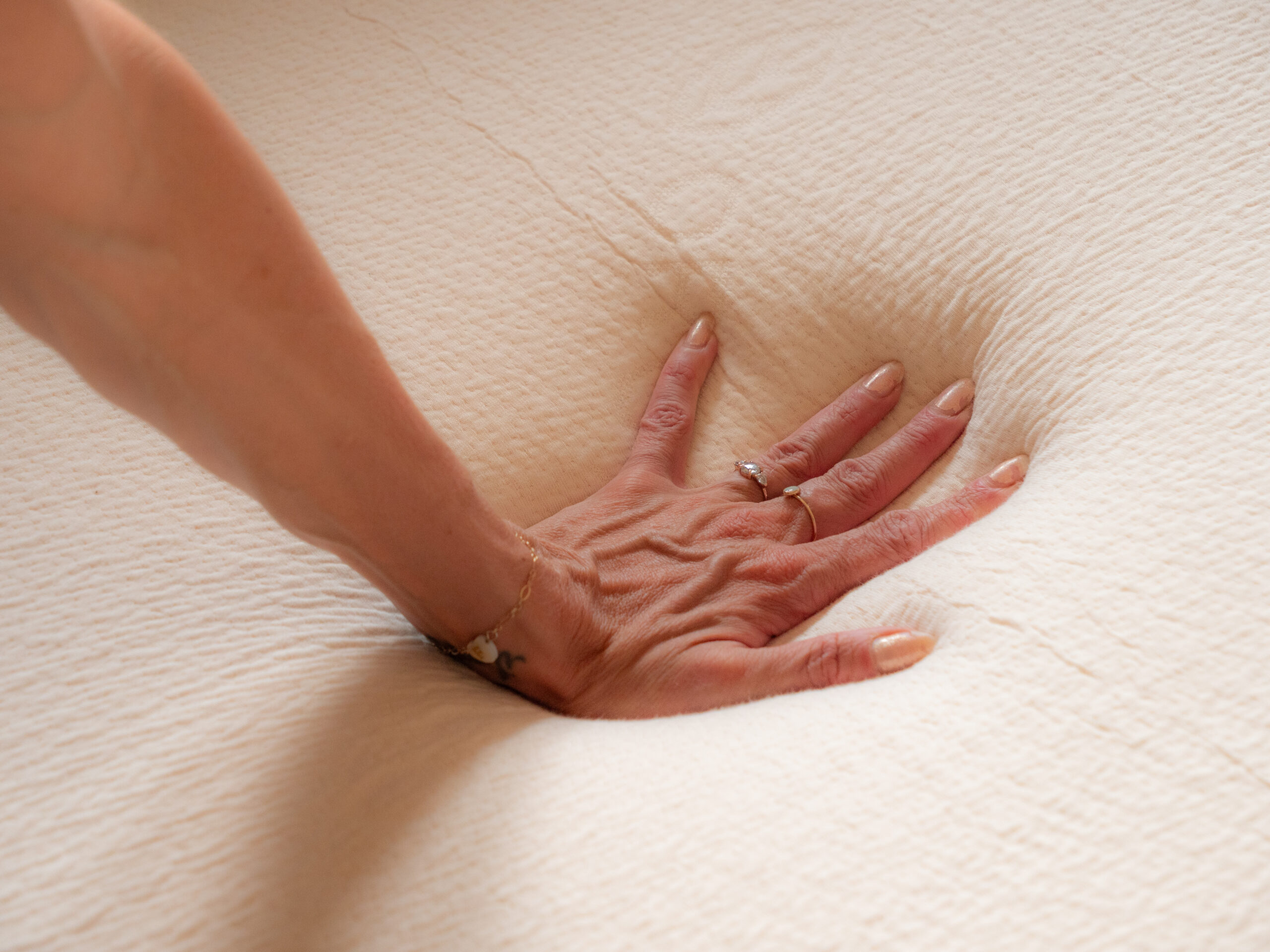 A hand presses down on a soft, beige mattress, creating a visible impression in the material.