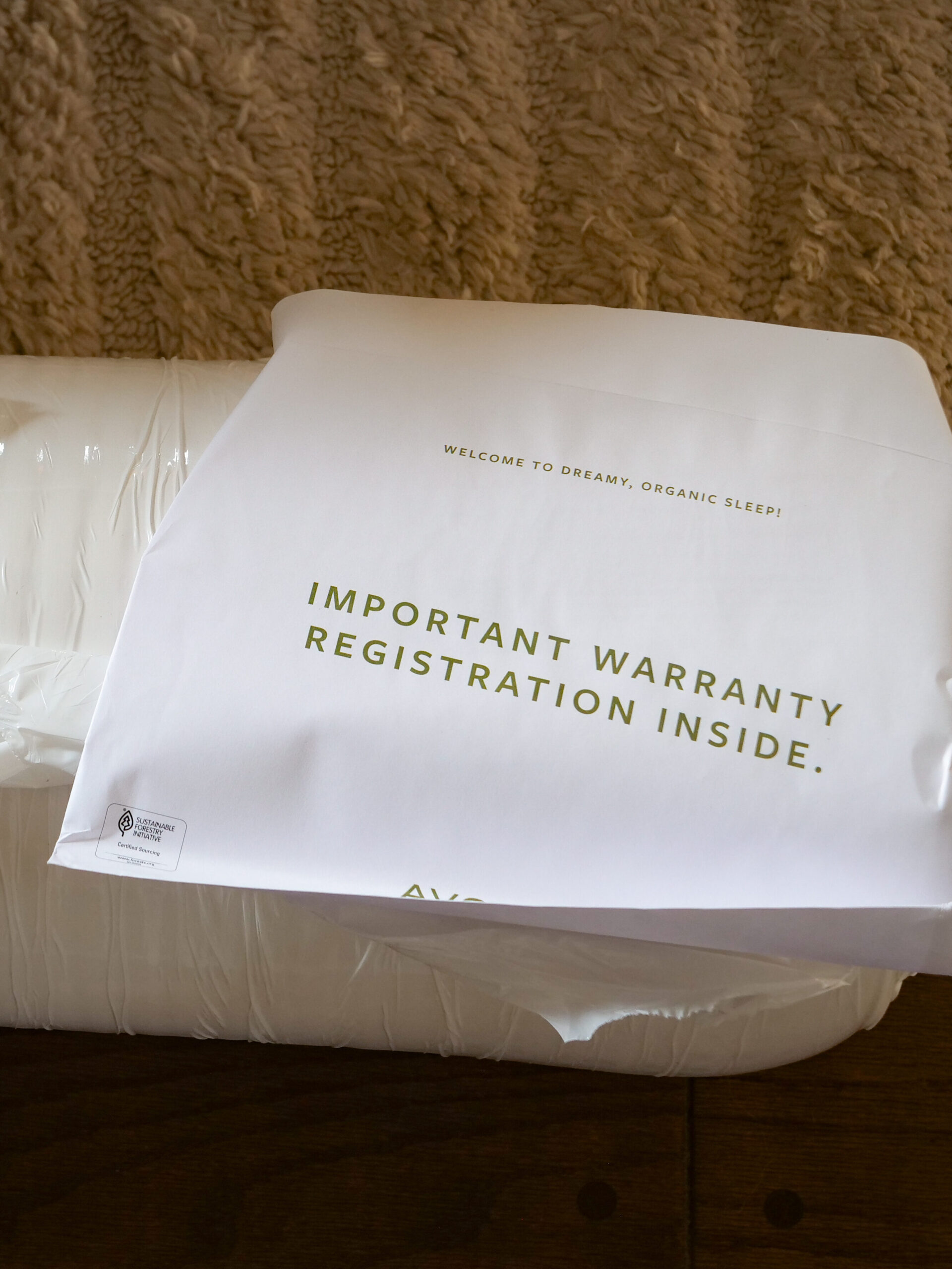 A mattress wrapped in plastic sits on a wooden floor with an Avocado booklet and a warranty registration envelope placed on top.