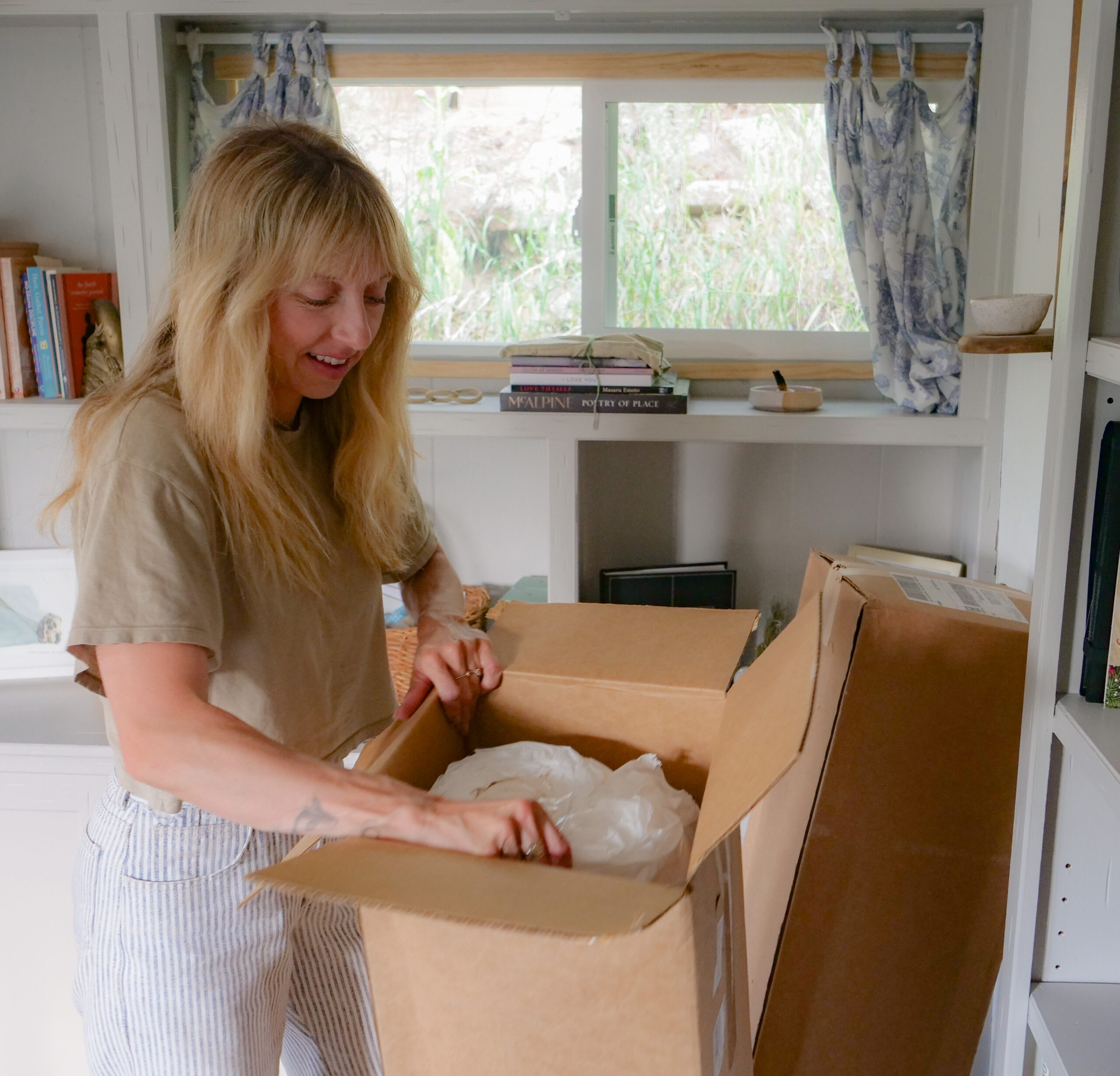 A woman stands in a room opening a cardboard box on a table, with shelves and a window in the background.