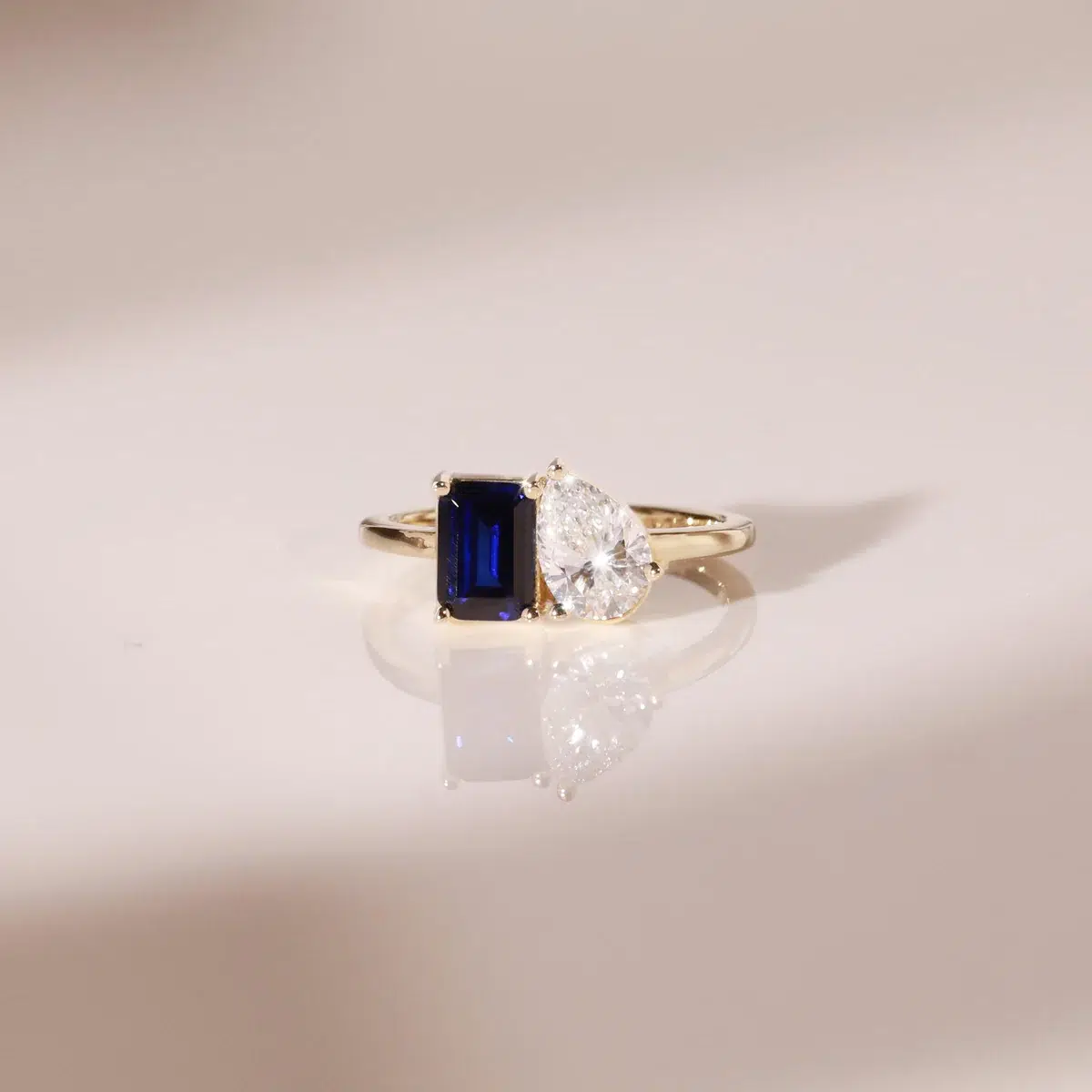 A gold ring featuring a rectangular blue gemstone set next to a pear-shaped clear gemstone on a smooth, light-colored surface.