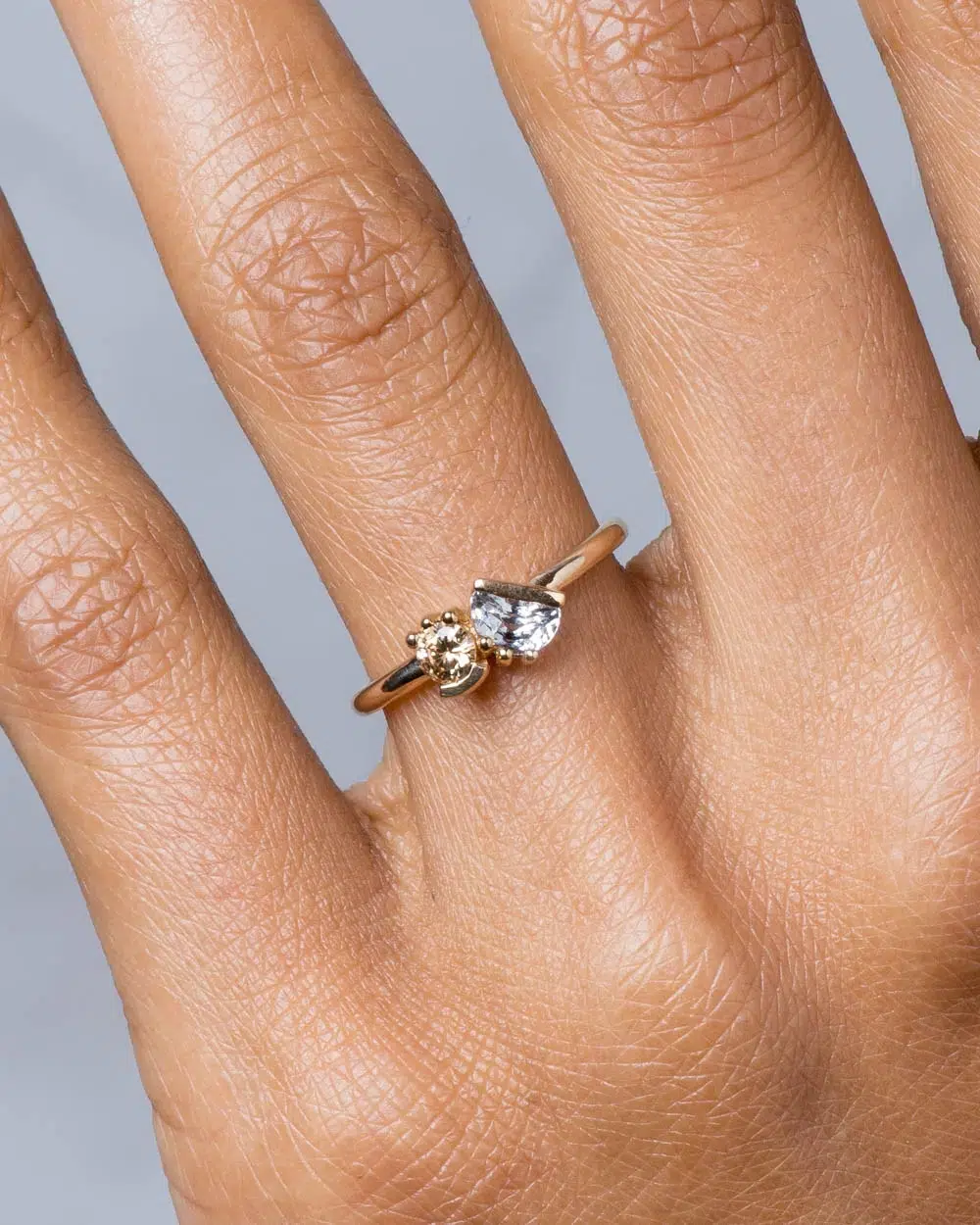 A hand wearing a gold ring featuring two asymmetrical gemstones, one clear and one gold, on the ring finger against a neutral background.