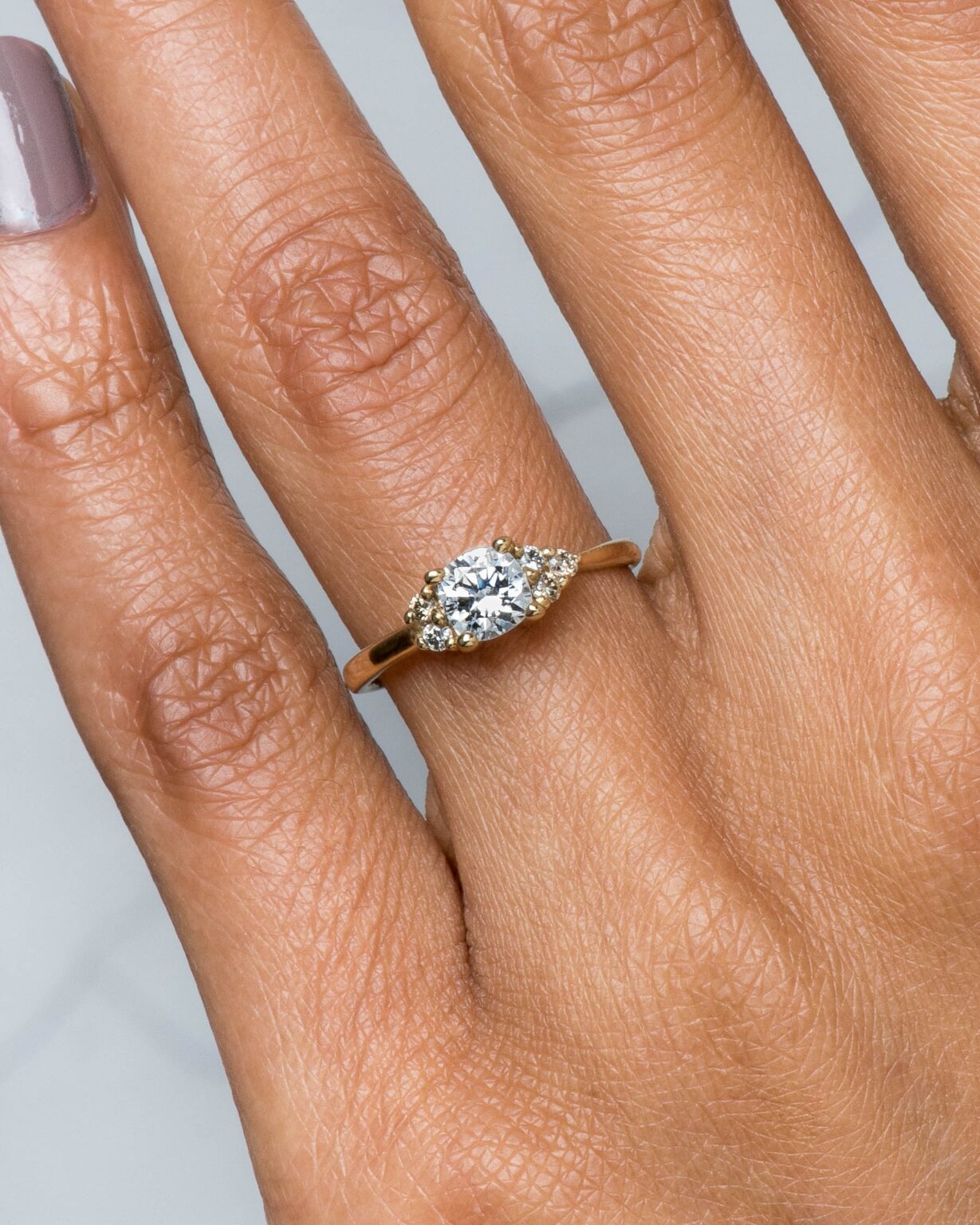 A hand with neutral-colored nail polish wearing a gold ring featuring a marquise-cut diamond flanked by two smaller round diamonds.