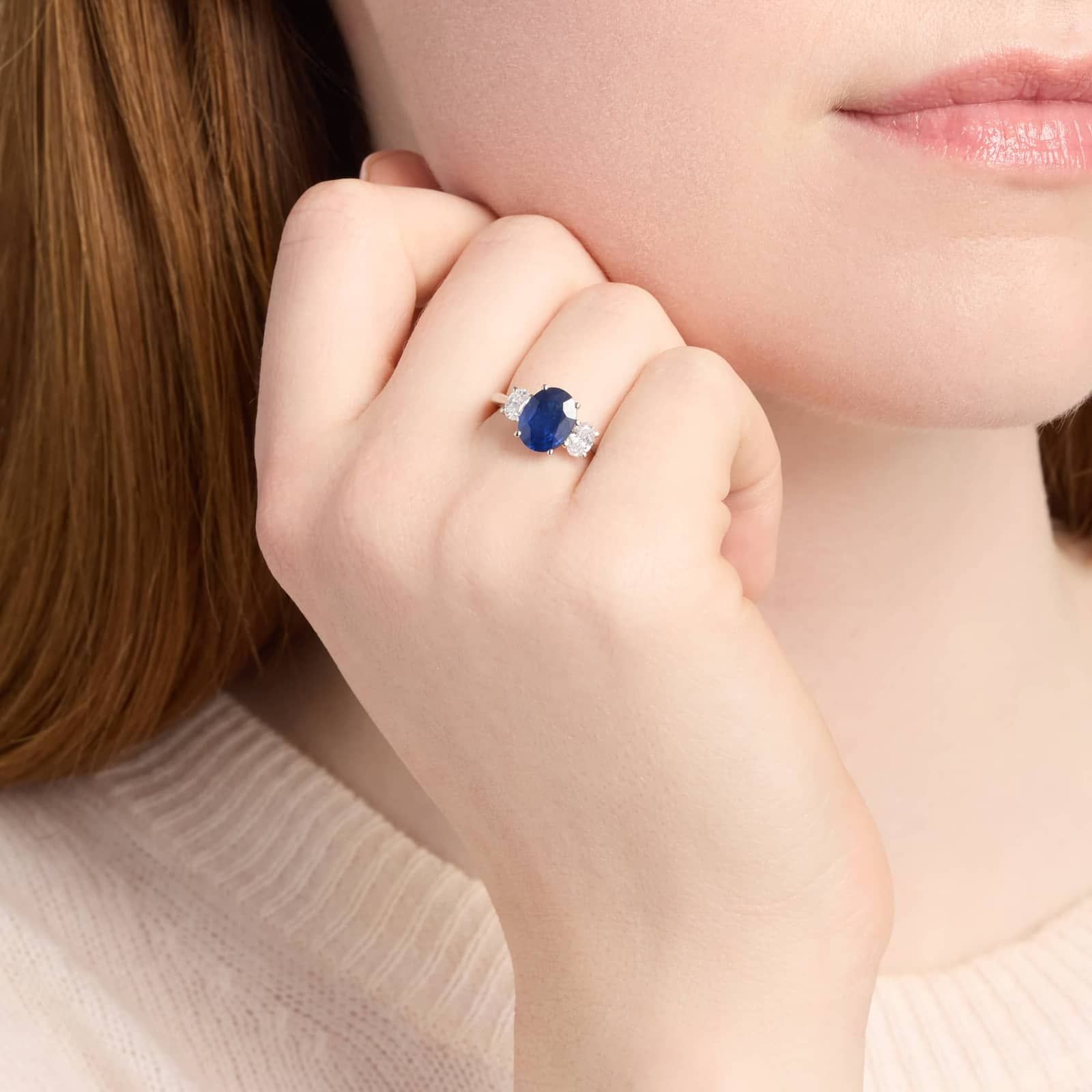 A person wears a silver ring with a large oval blue gemstone flanked by two clear stones on the ring finger of their left hand.