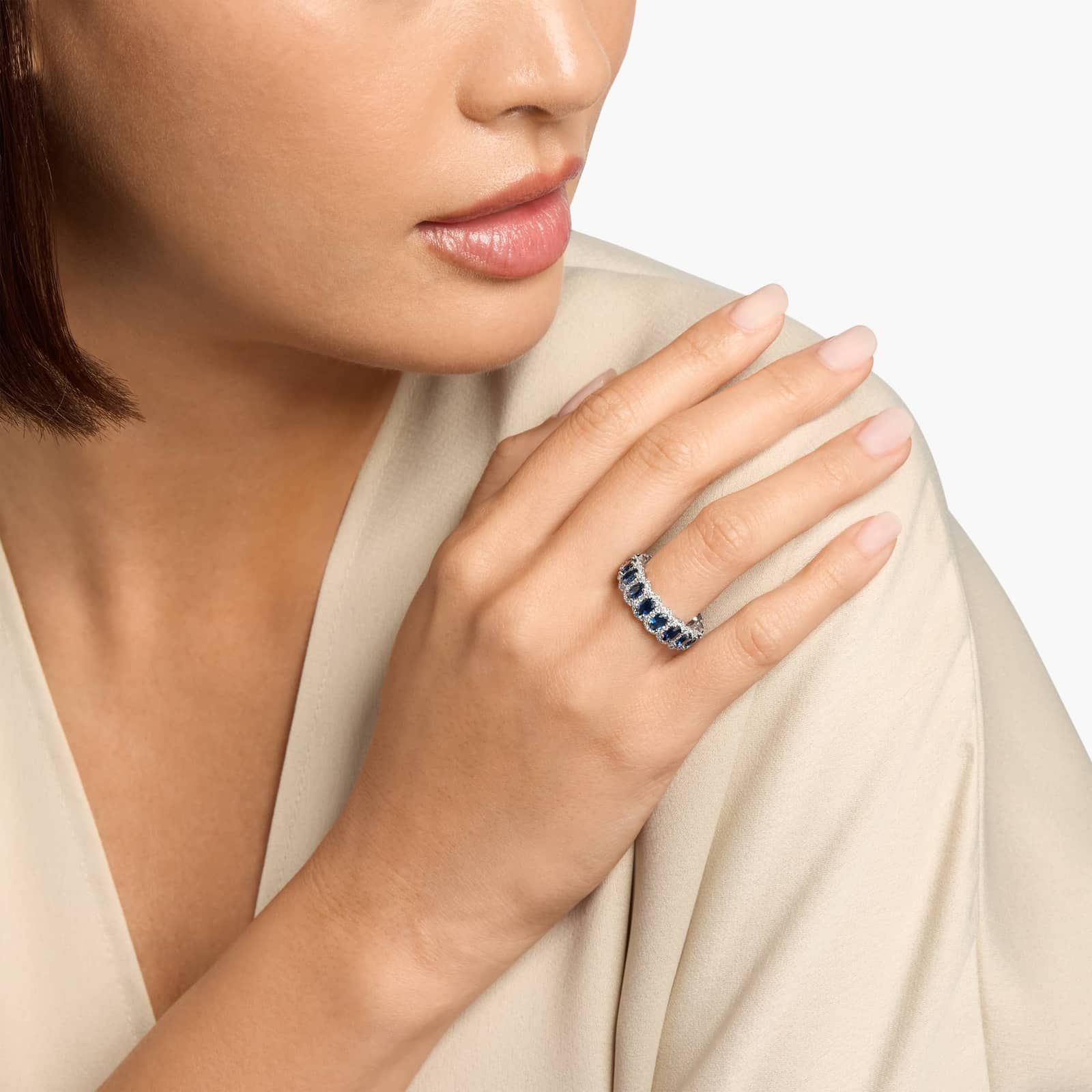 A woman in a beige top rests her hand on her shoulder, showcasing a silver ring with blue stones on her finger.