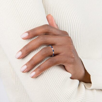 A hand with neatly manicured nails rests on an arm, wearing a thin silver ring with blue stones, against a white ribbed long-sleeve top.