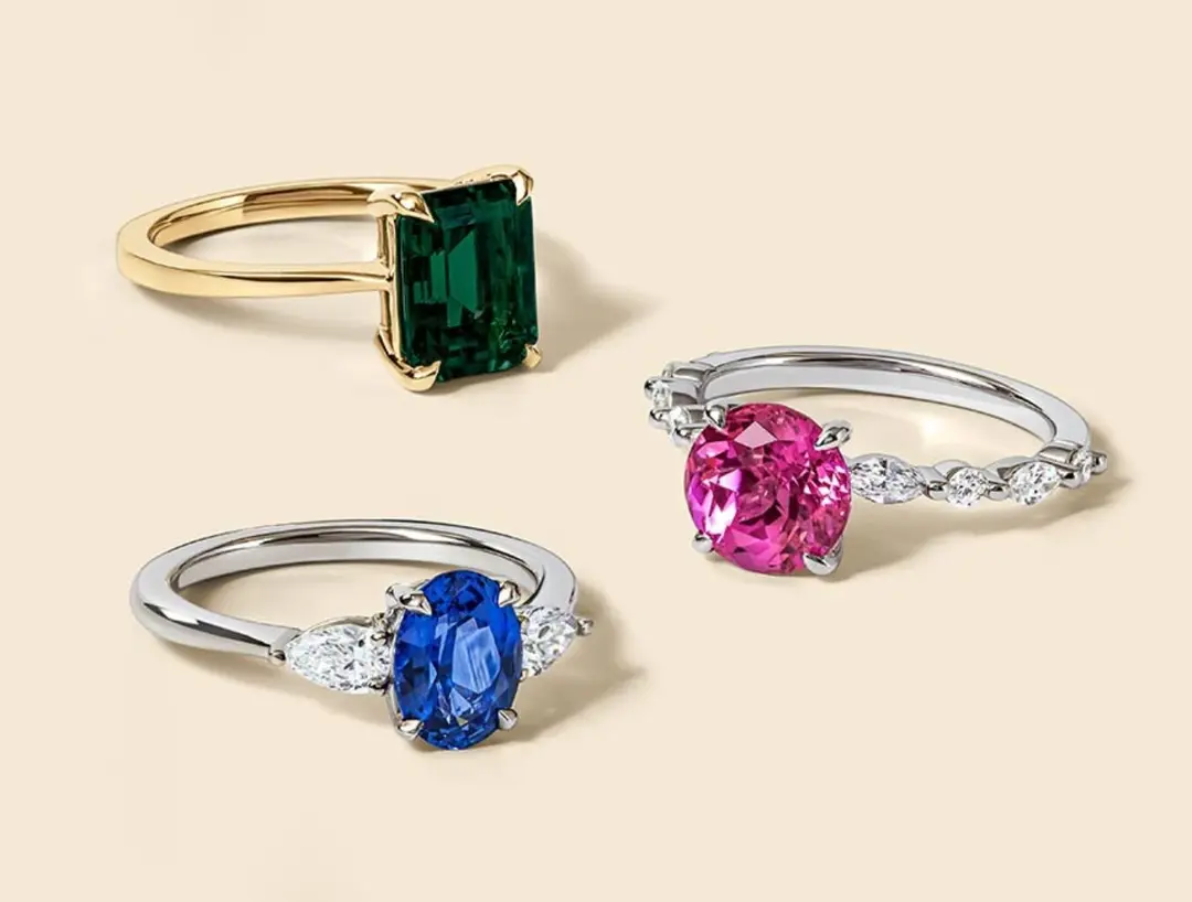 Three gemstone rings: one with a rectangular green stone on a gold band, one with an oval pink stone and two small clear stones, and one with an oval blue stone and two small clear stones.