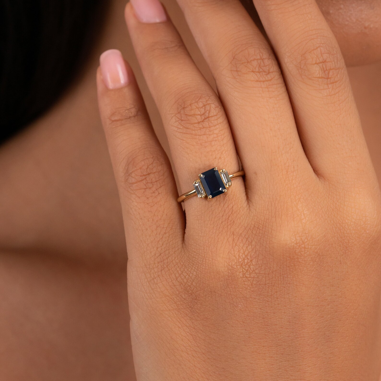 A hand with neatly manicured nails wears a gold ring featuring a rectangular dark blue gemstone flanked by small clear stones.