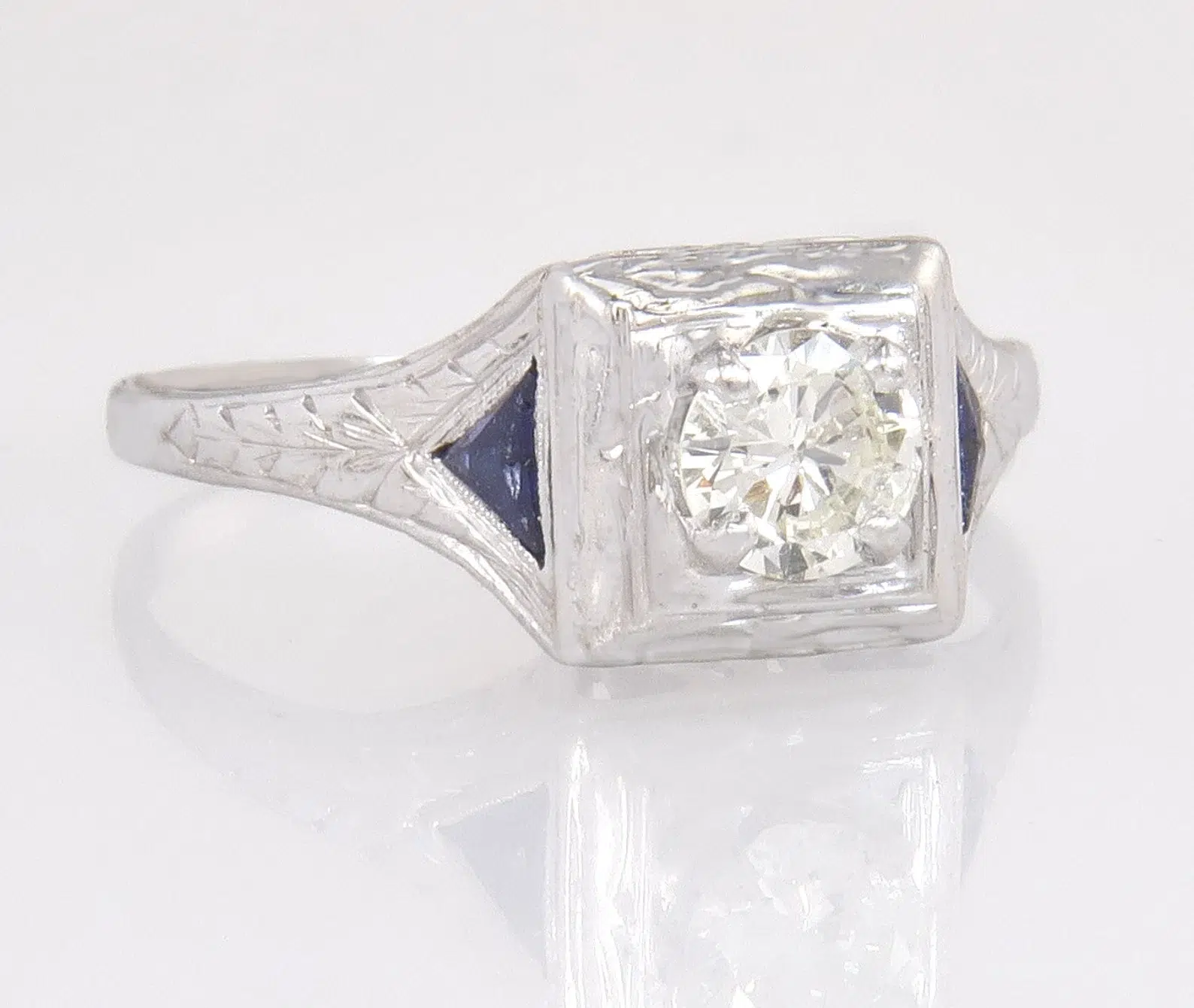 A silver ring with an engraved band features a round, clear gemstone set in a square bezel, flanked by small dark blue triangular accents.