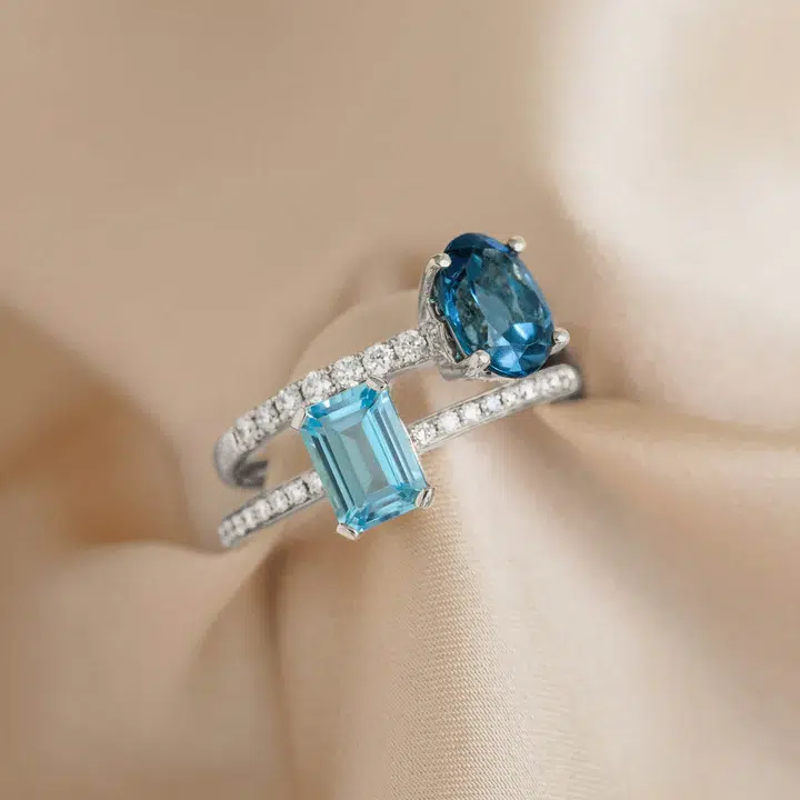 A silver ring with two blue gemstones—one oval and one rectangular—set on a band with small clear stones, displayed on a beige satin fabric.