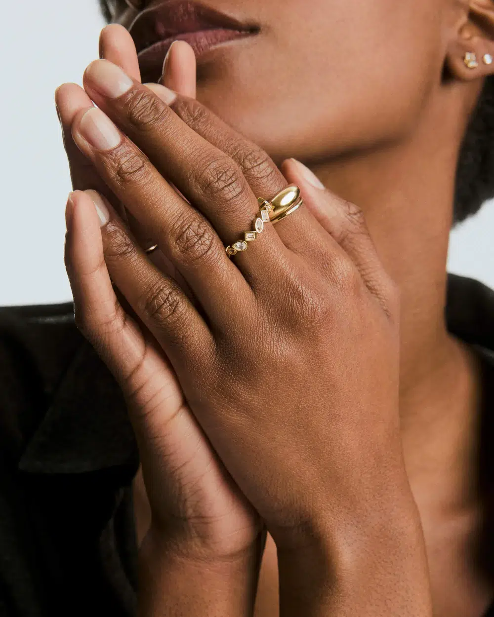 A person with dark skin holds their hands near their face, showcasing two gold rings on their fingers. The background is neutral, and the clothing is black.