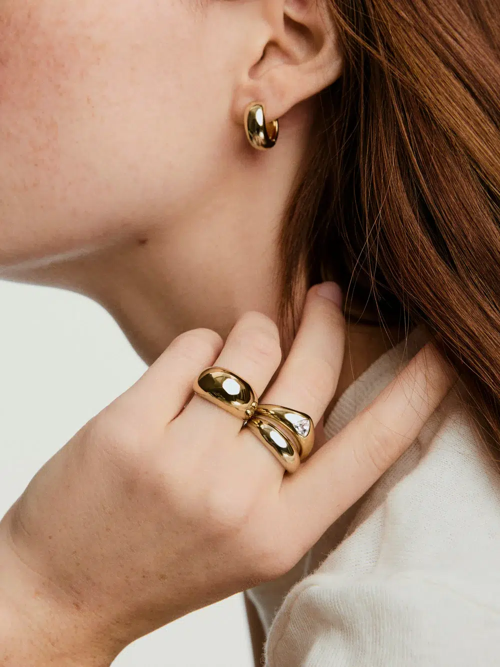 A person wearing a gold hoop earring and two chunky gold rings on their fingers, with their hand resting near their face.