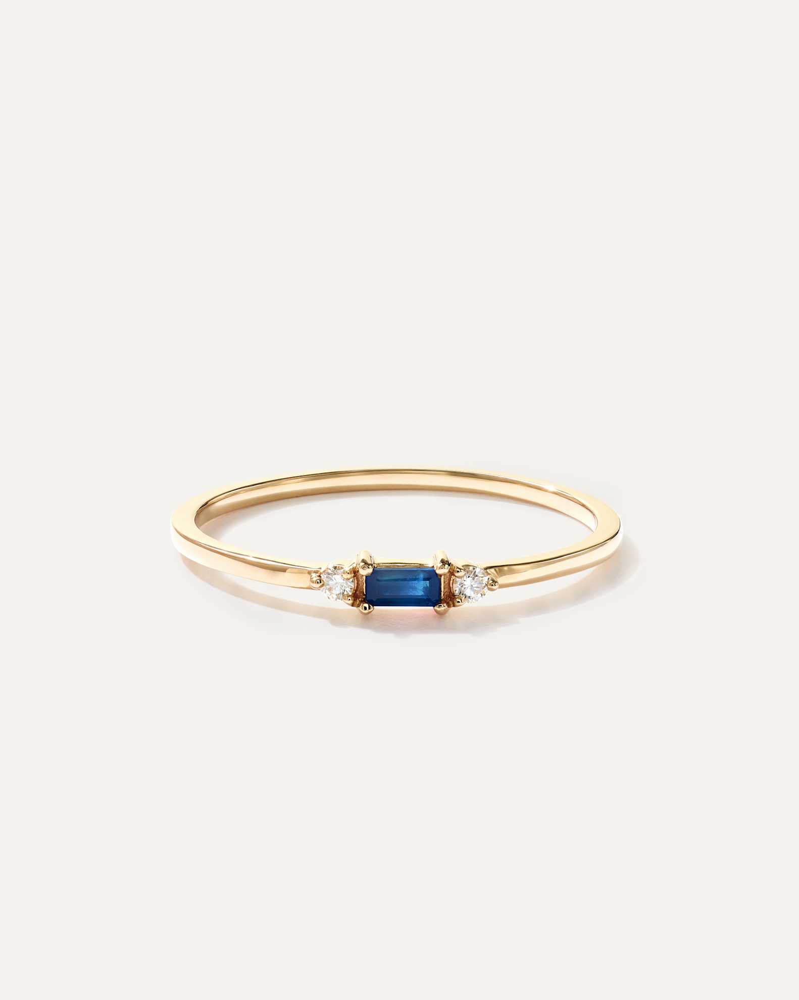 A thin gold ring with a rectangular blue gemstone at the center, flanked by two small round clear stones, displayed on a plain light background.