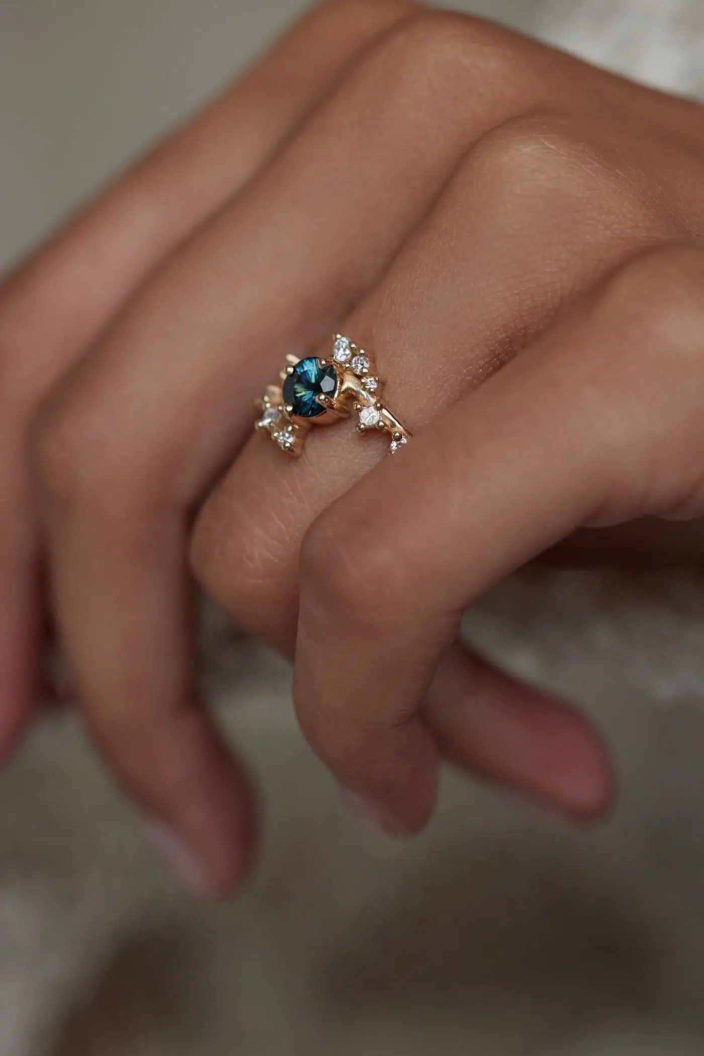 A hand wearing a gold ring with a central blue gemstone surrounded by small clear stones, set in an asymmetrical design.