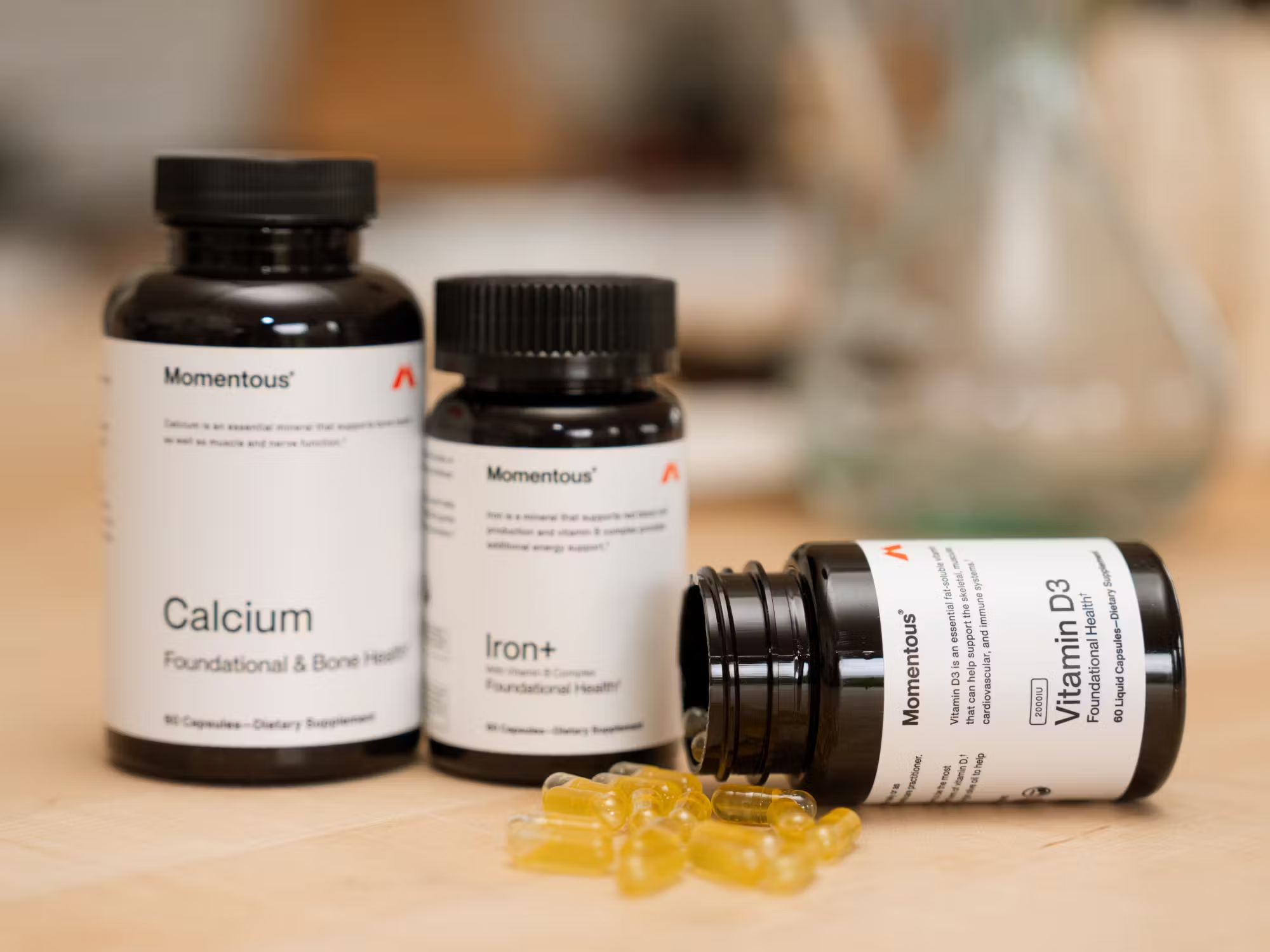 Three bottles of Momentous dietary supplements labeled Calcium, Iron+, and Vitamin D3 sit on a table, with several yellow capsules spilled in front of the Vitamin D3 bottle.