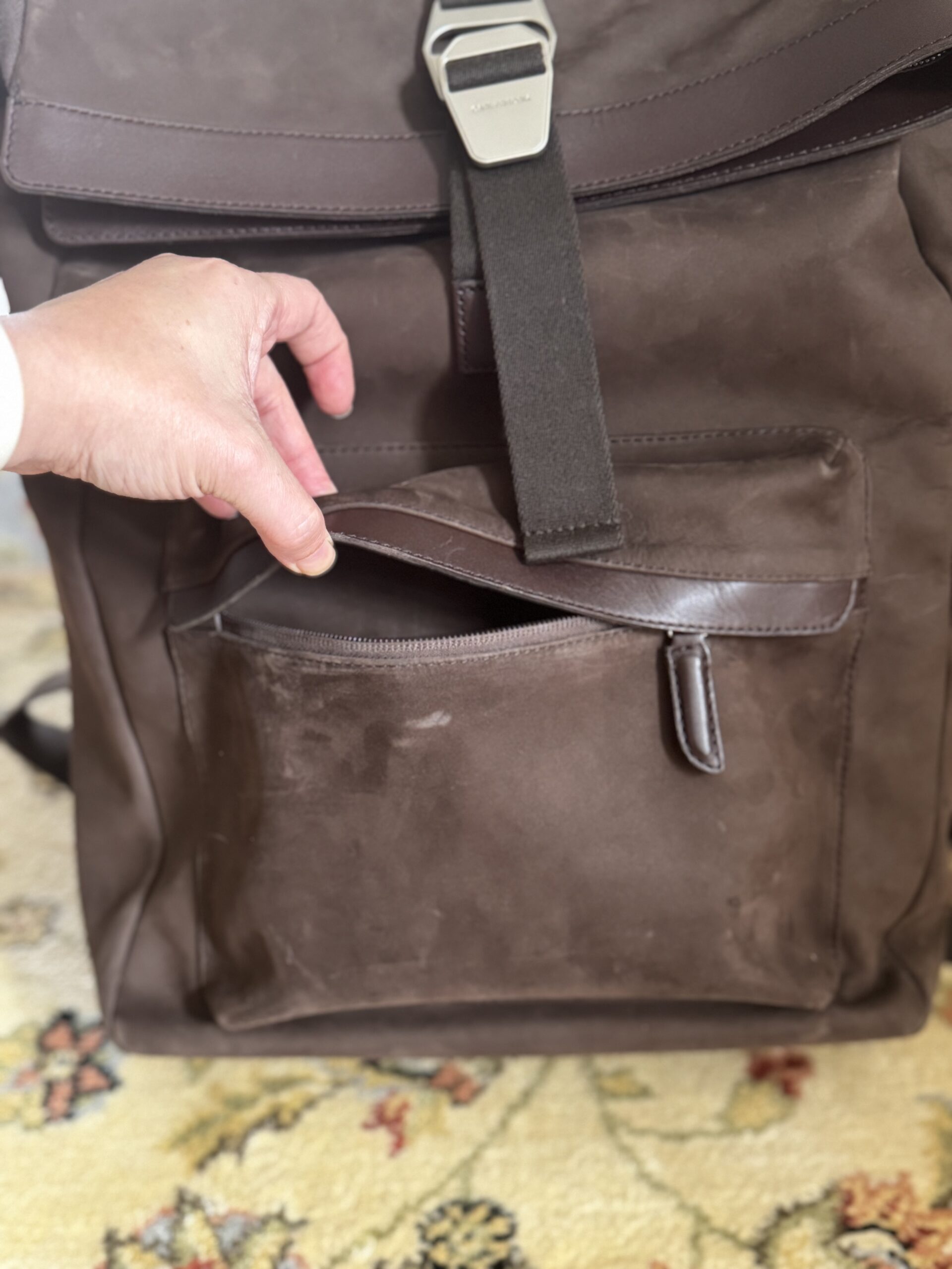 A hand is opening the front zipper pocket of a brown leather backpack resting on a patterned carpet.