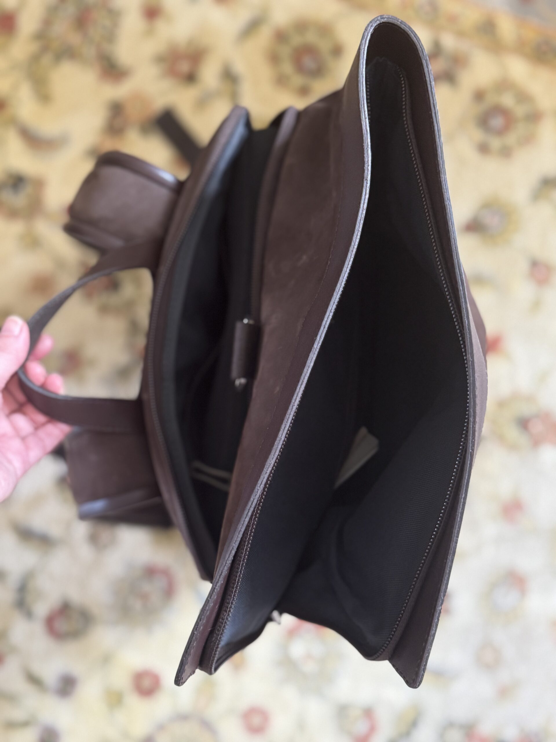 A hand holds open a brown leather bag, revealing its spacious black interior and side pocket, with a patterned carpet in the background.