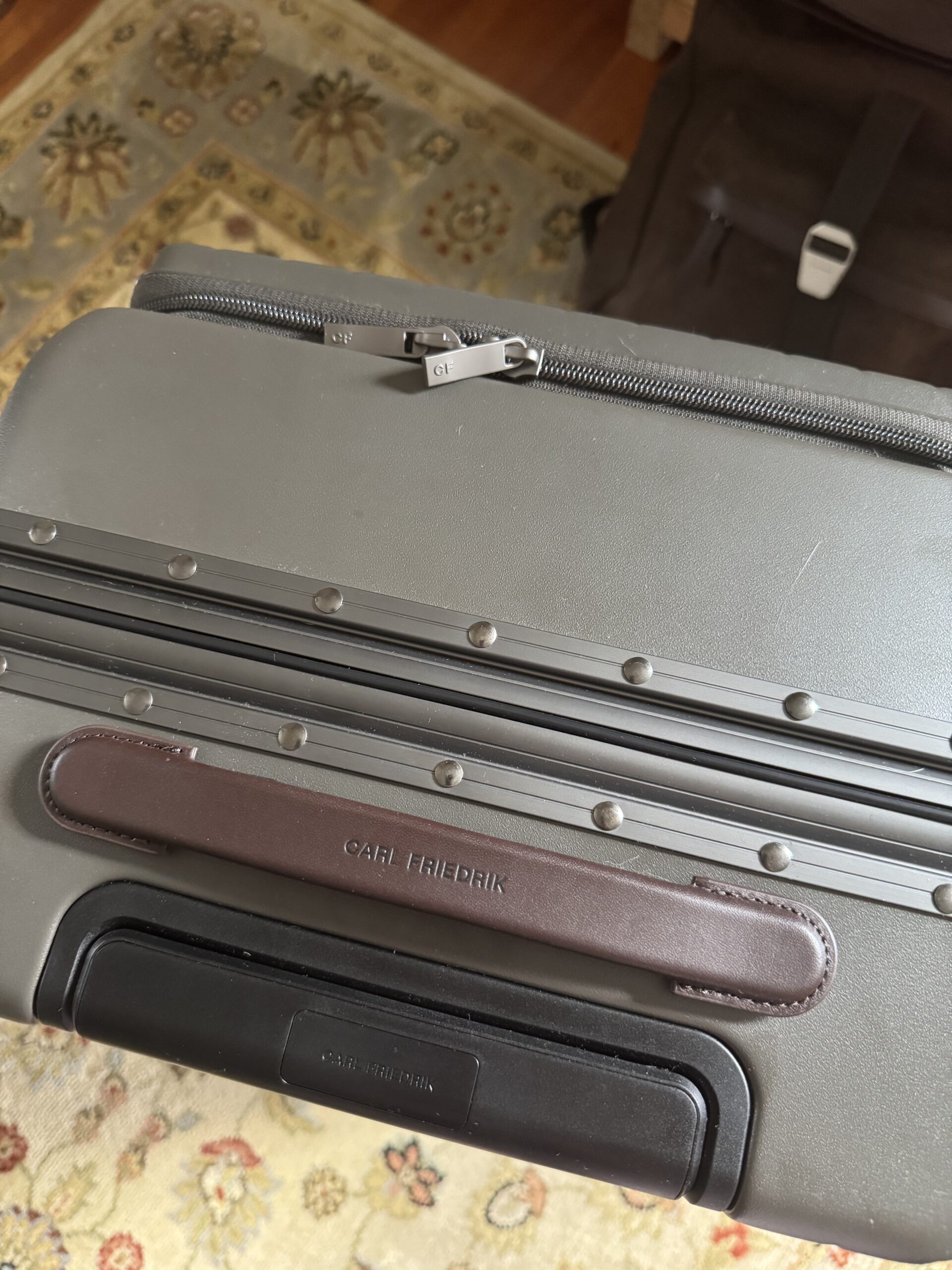 A close-up of a gray hard-shell suitcase with a brown leather handle labeled "Carl Friedrik" on a patterned rug.