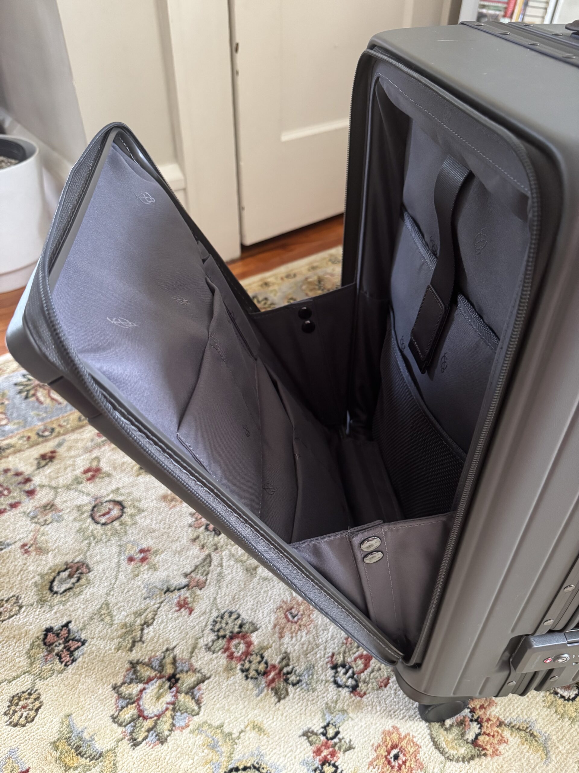 Gray hard-shell suitcase with its front compartment open, showing the empty interior. The suitcase is on a patterned carpet near a white door.