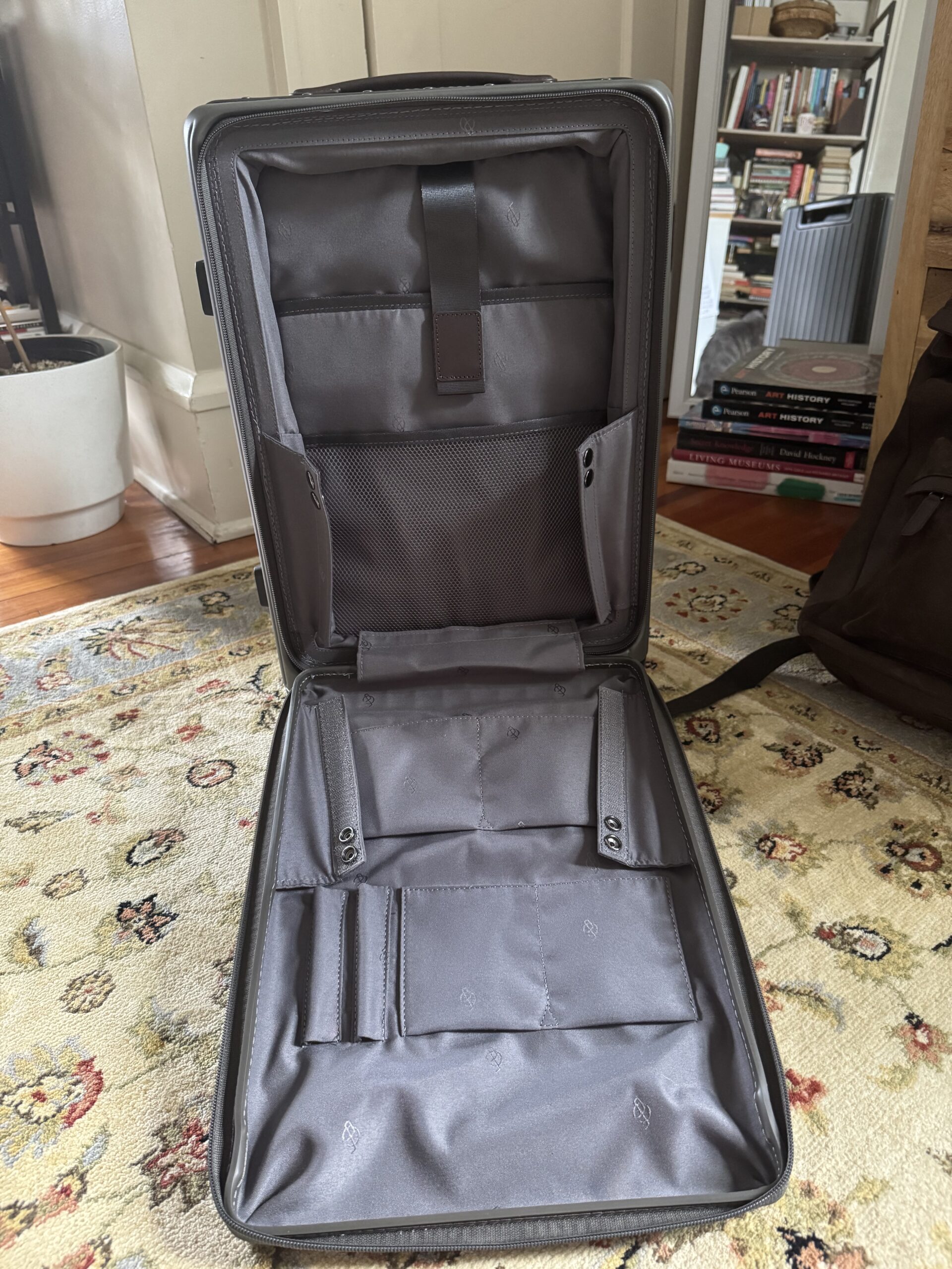 Open, empty gray suitcase with organized interior pockets and straps, placed on a patterned rug in a room with bookshelves and a plant in the background.