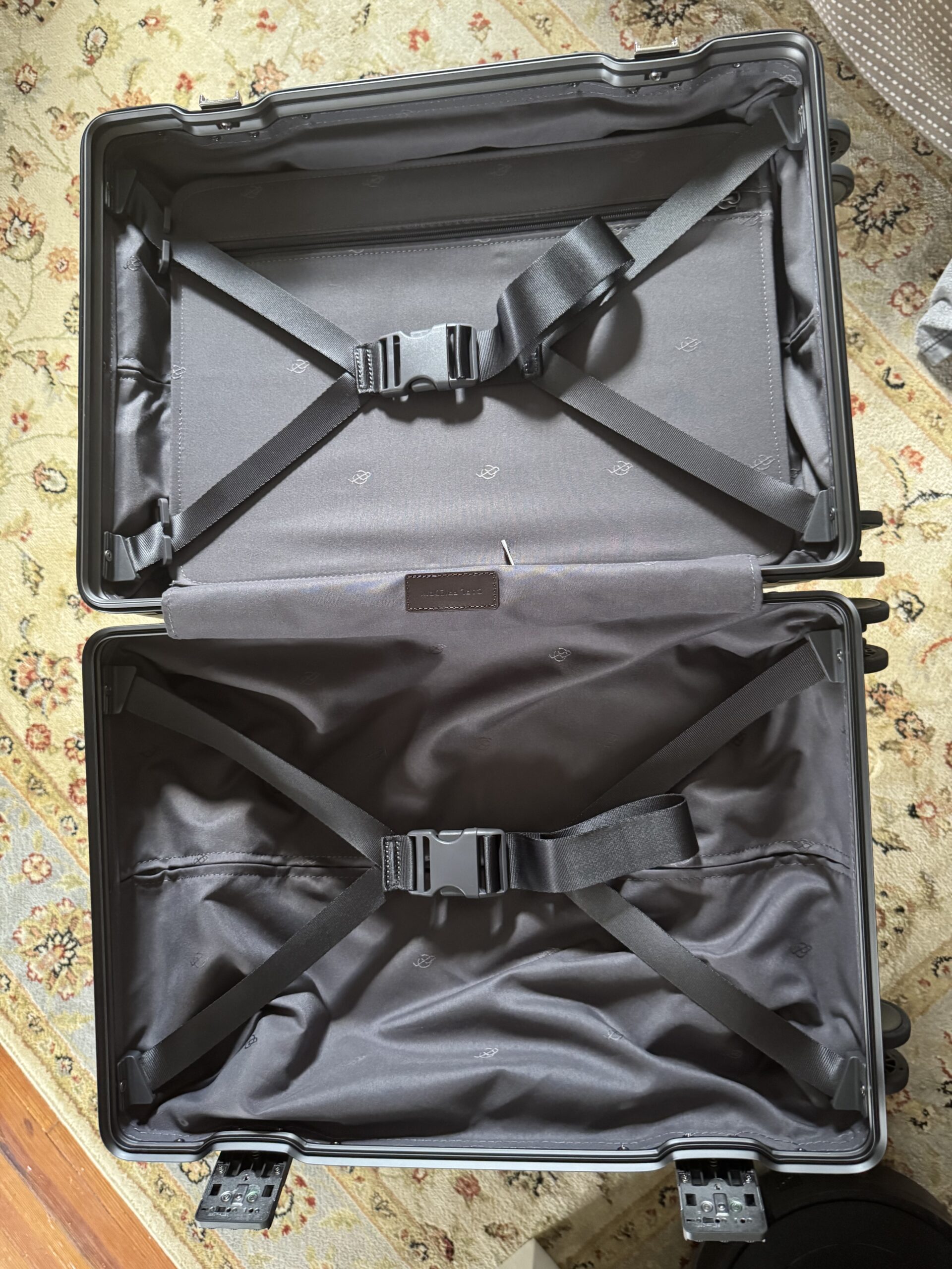 Open black hard-shell suitcase with two compartments, each secured by adjustable cross straps, placed on a patterned carpet.