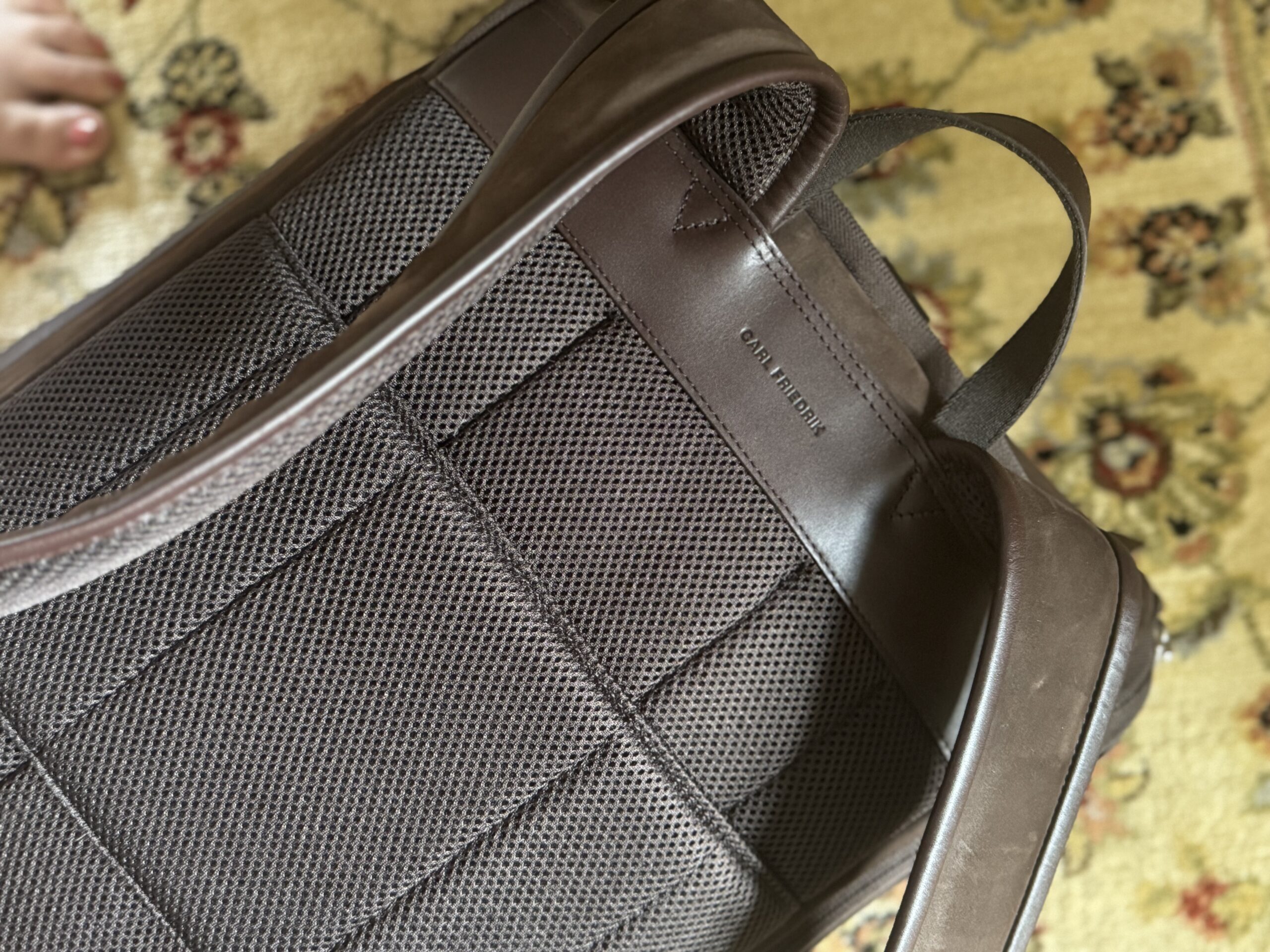 A close-up of the back side of a brown leather backpack with mesh padding, placed on a patterned carpet. A person's toes are visible at the top left corner.
