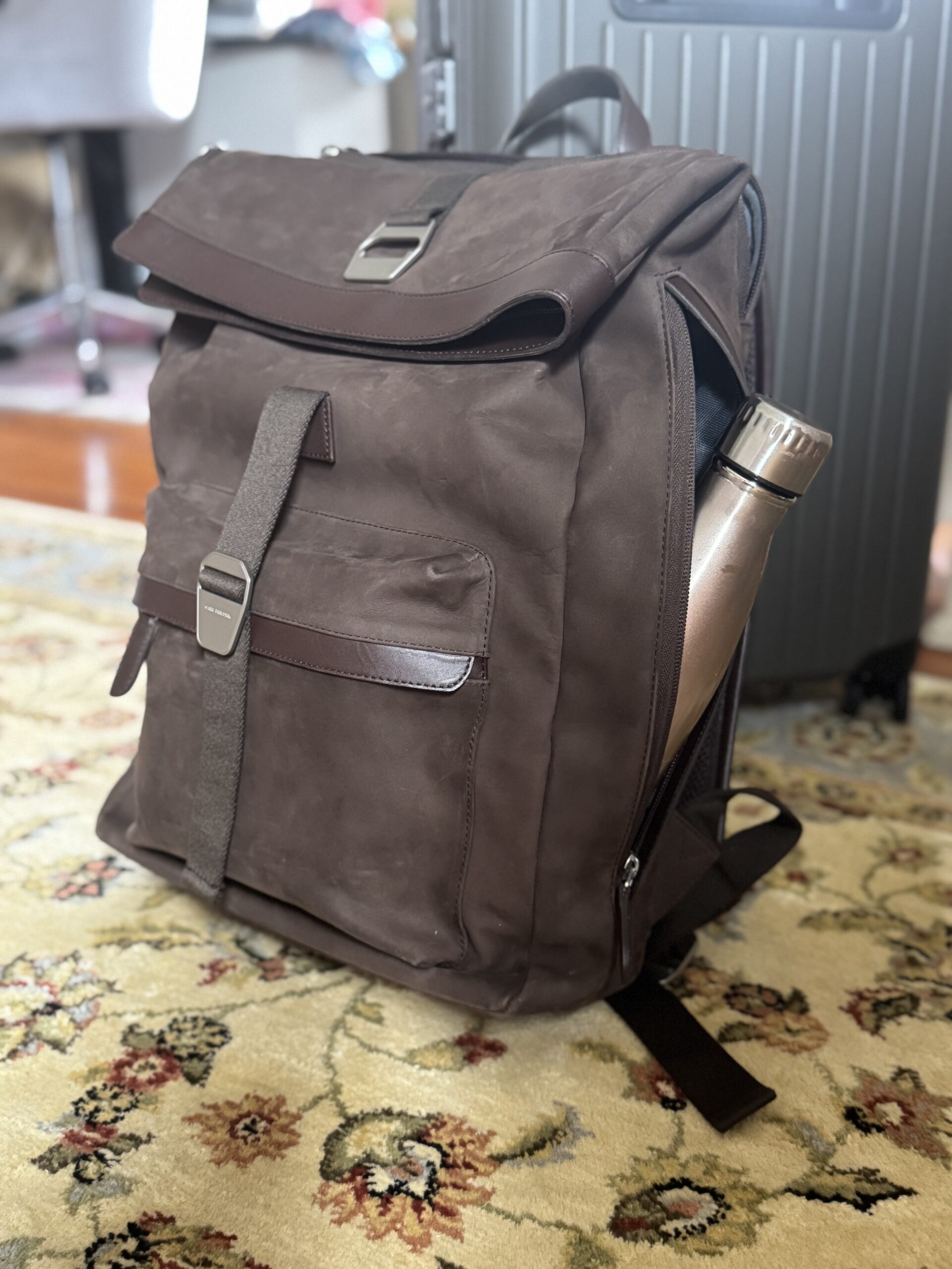 A brown backpack with a front pocket and metal buckle sits on a floral rug, partially unzipped to reveal a silver water bottle inside.