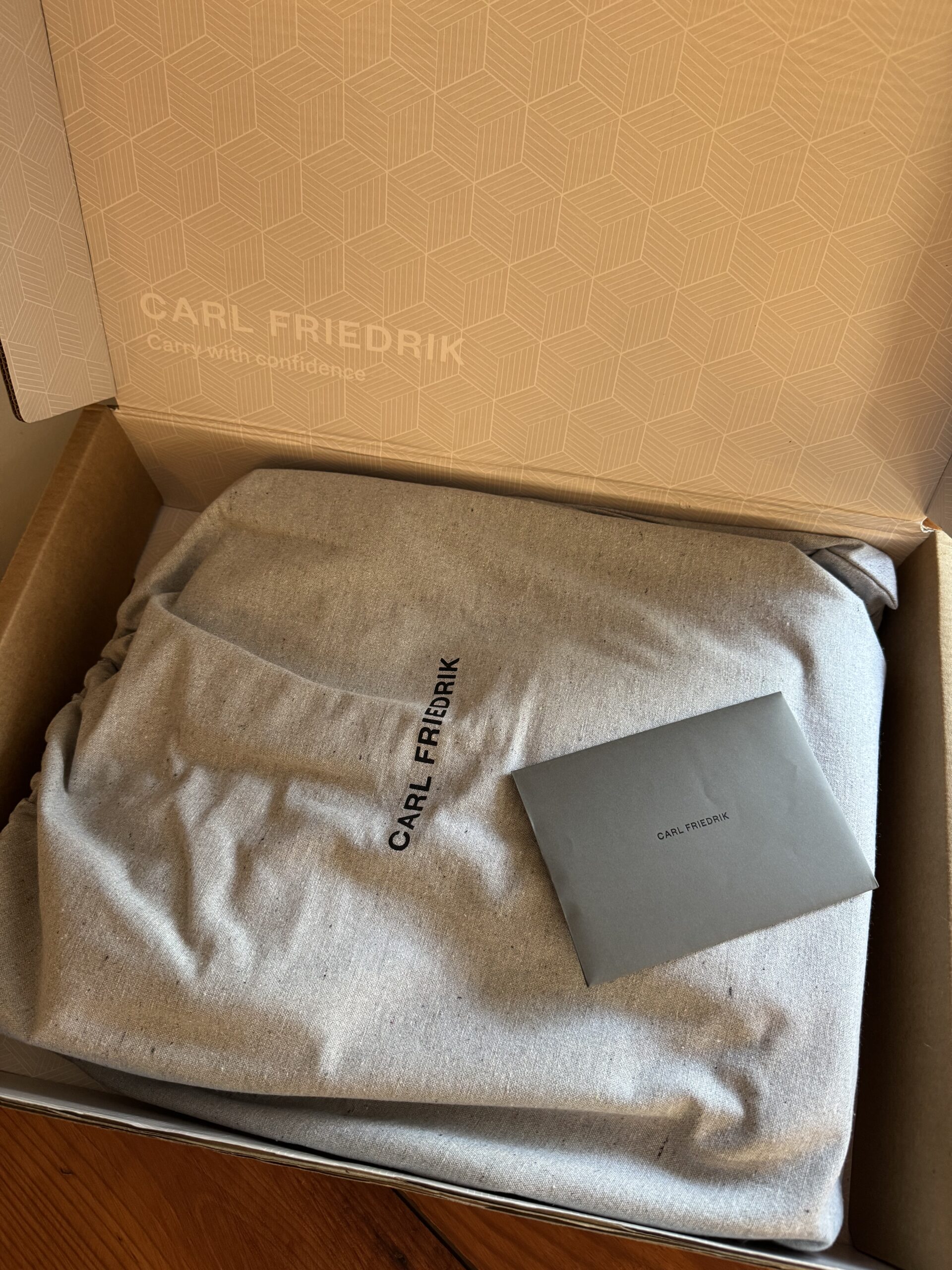 A Carl Friedrik-branded item covered in a grey dust bag is placed inside an open box, accompanied by a small grey envelope.