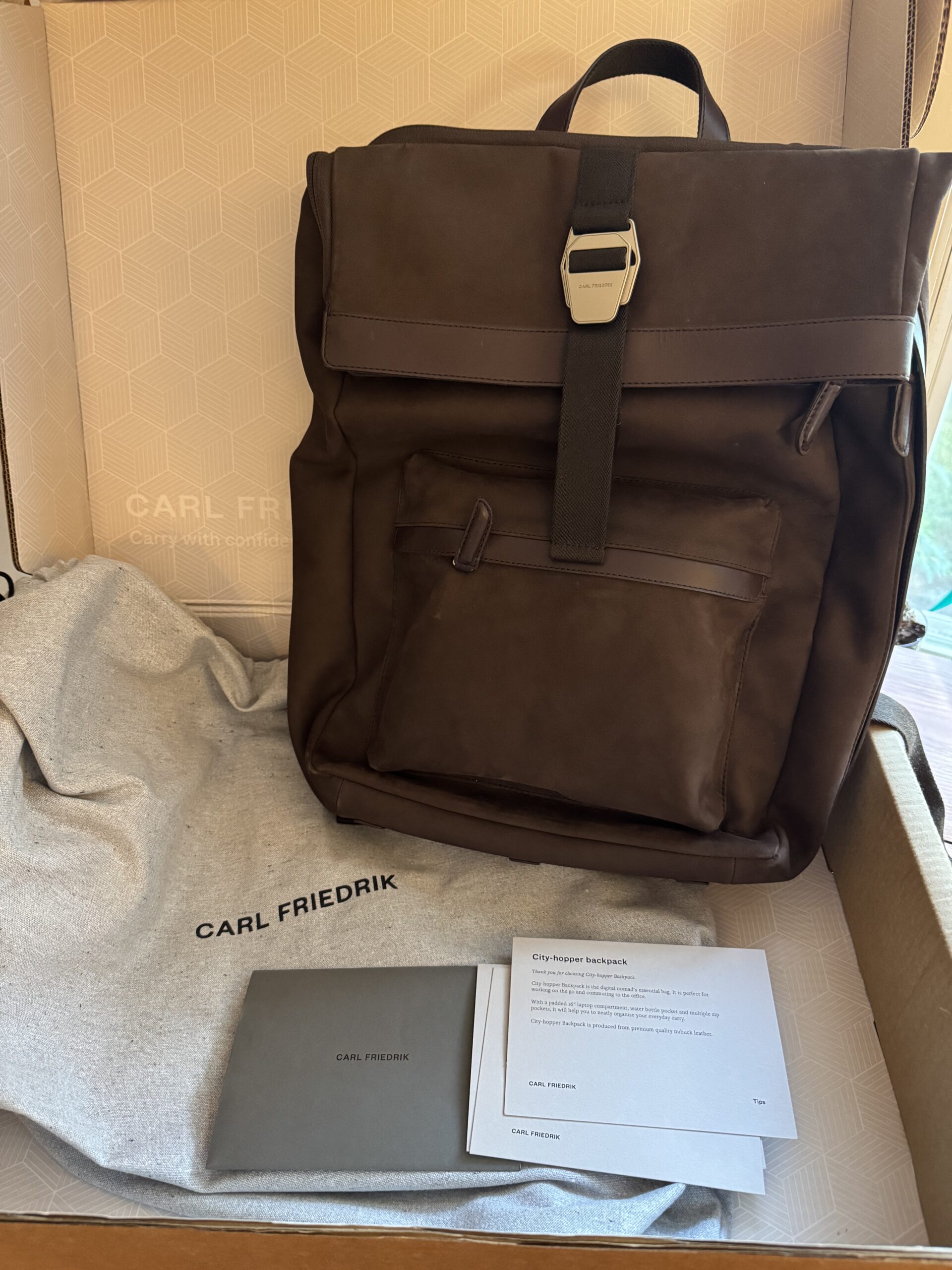 A brown Carl Friedrik backpack sits in an open box with a dust bag, an envelope, and an information card.