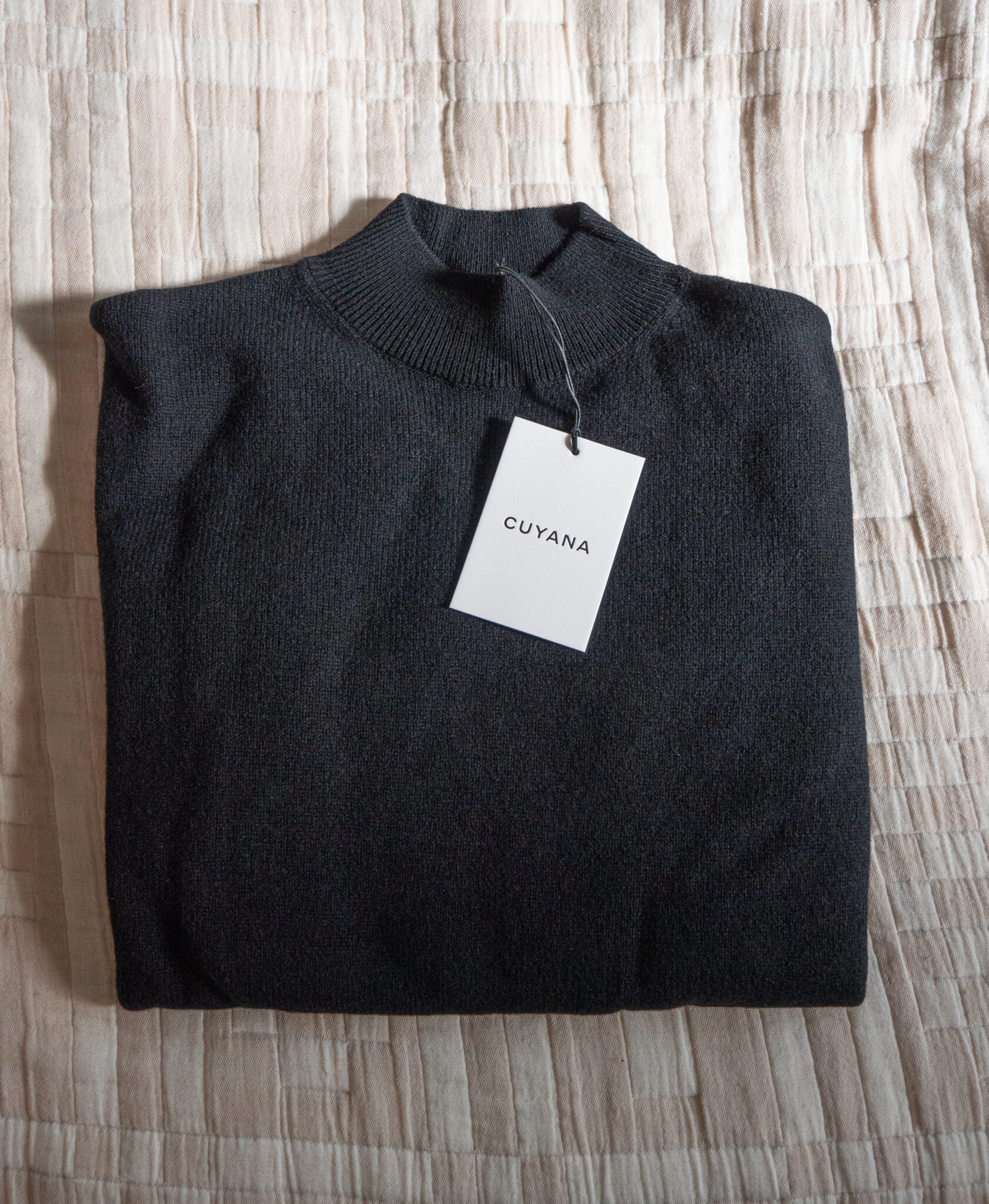 A folded black sweater with a Cuyana tag rests on a beige, textured surface.