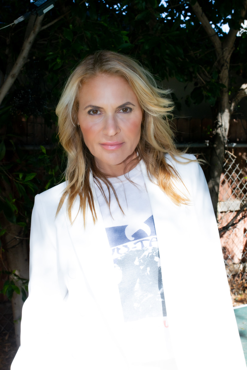 A woman with blonde hair wearing a white blazer and a printed T-shirt stands outdoors in partial sunlight near trees and a chain-link fence.