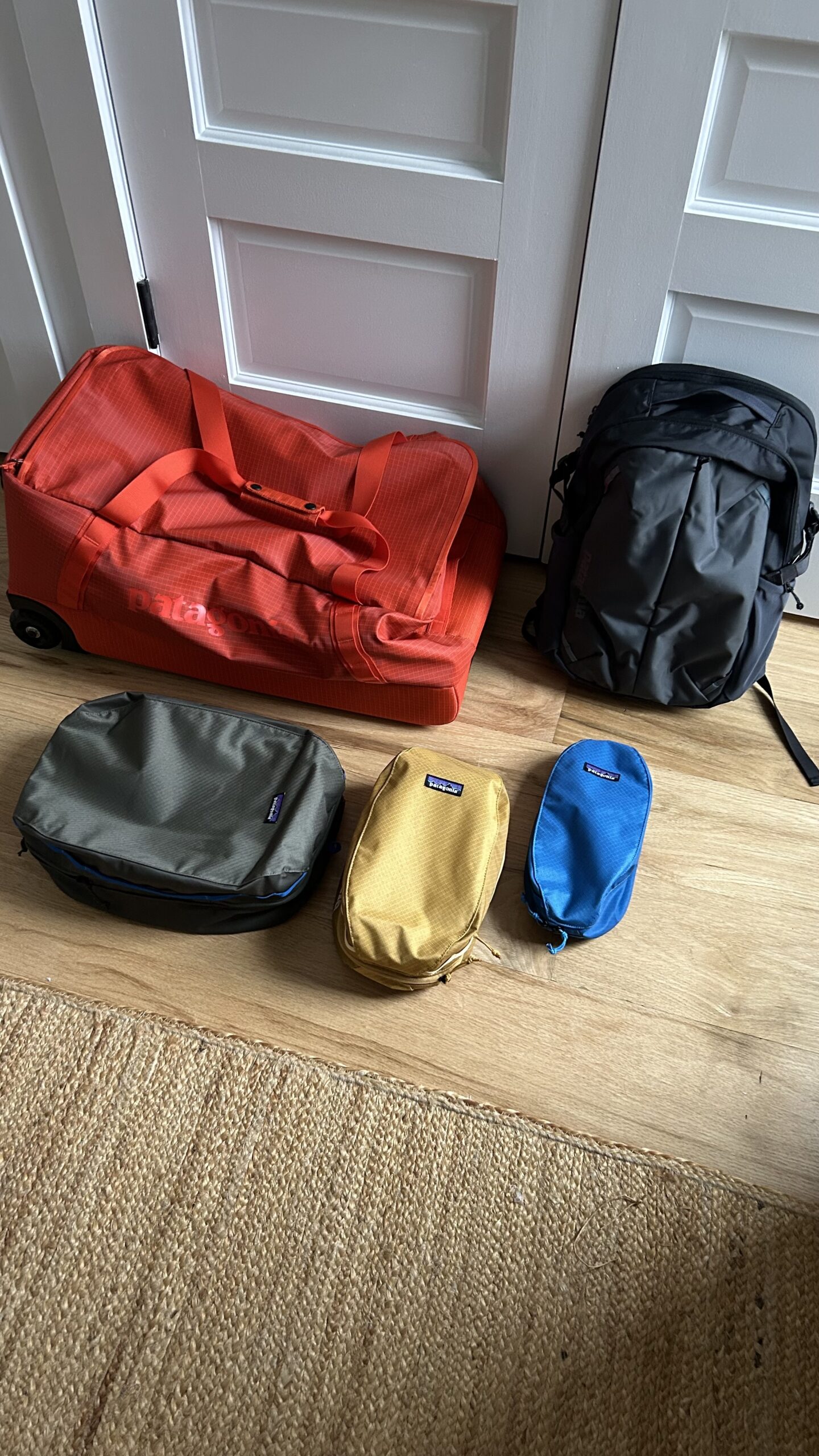 A red duffel bag, a black backpack, and three small zip pouches in green, yellow, and blue are arranged on a wooden floor in front of white double doors.