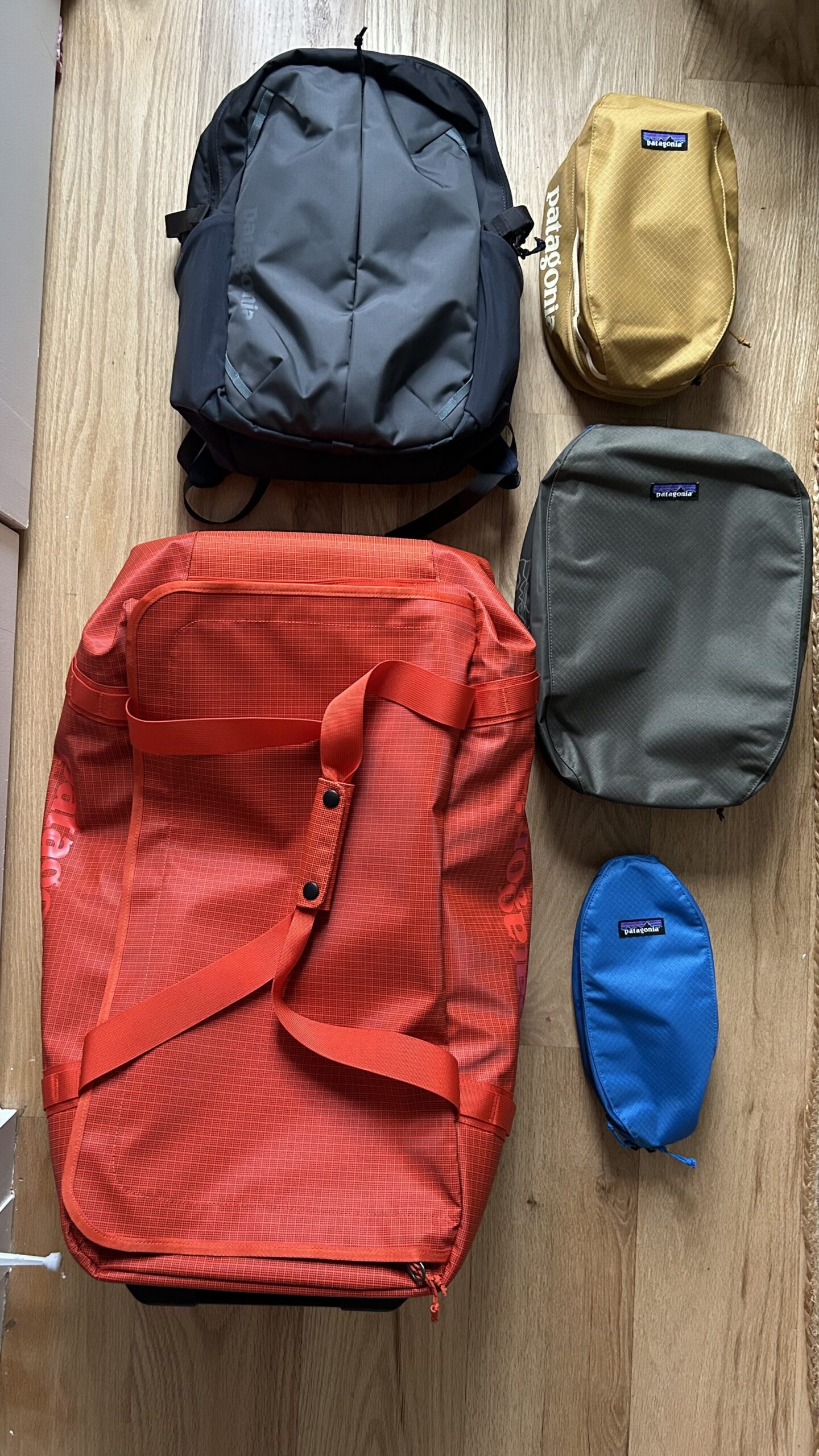 Five Patagonia travel bags of different sizes and colors are laid out on a wooden floor, including a large red duffel and four smaller packing cubes.