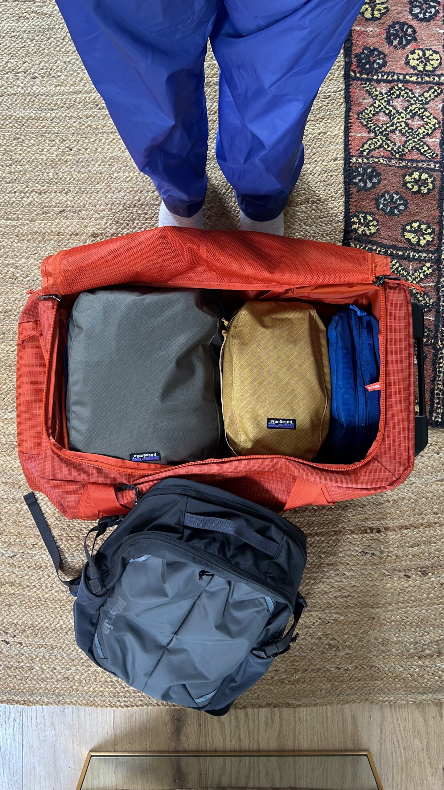A person in blue pants stands by an open red duffel bag packed with three packing cubes and a gray backpack on the floor.