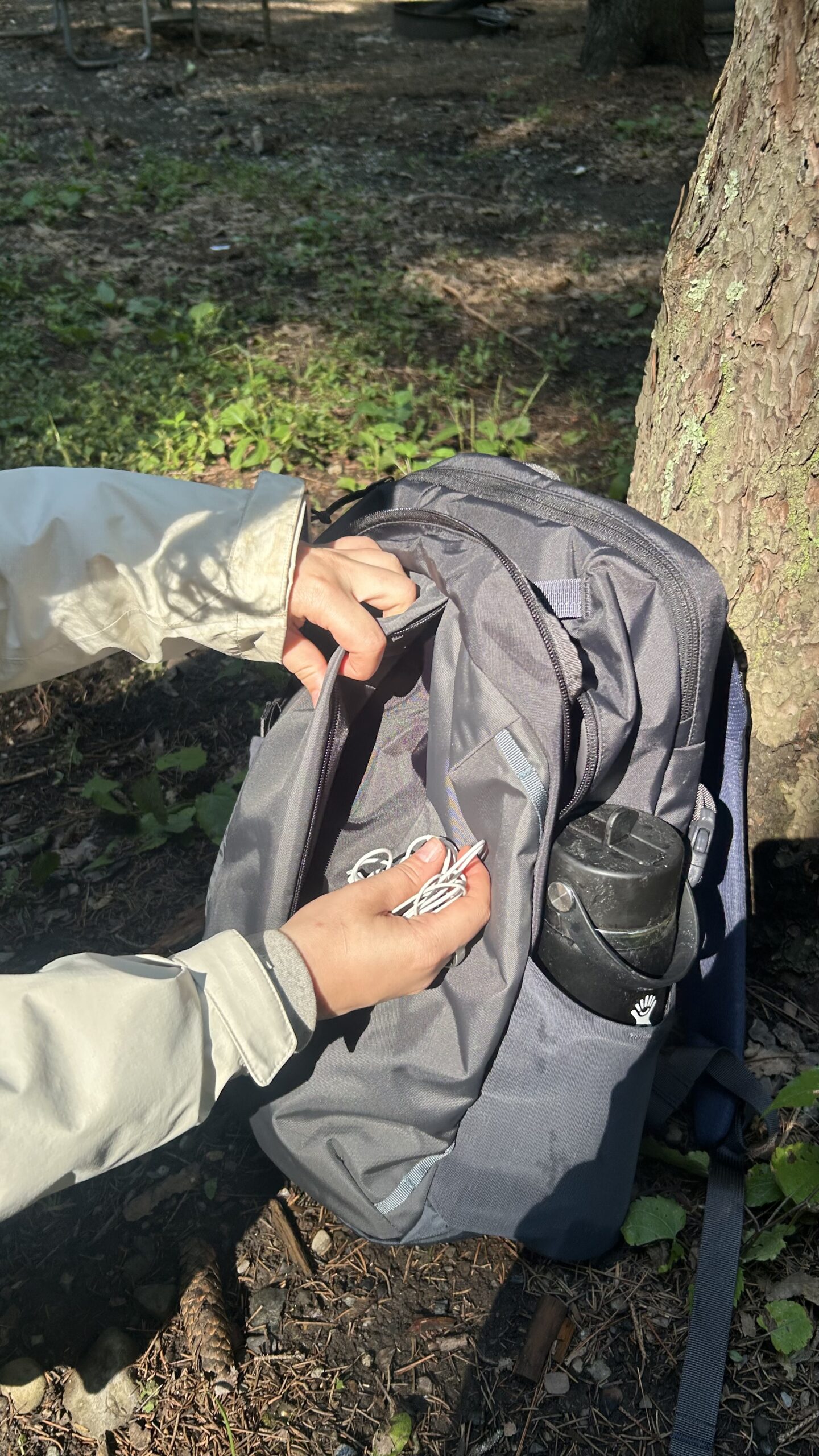 Person opening a gray backpack outdoors, holding earphones in one hand; a black water bottle is in the side pocket. Sunlight and trees are visible in the background.