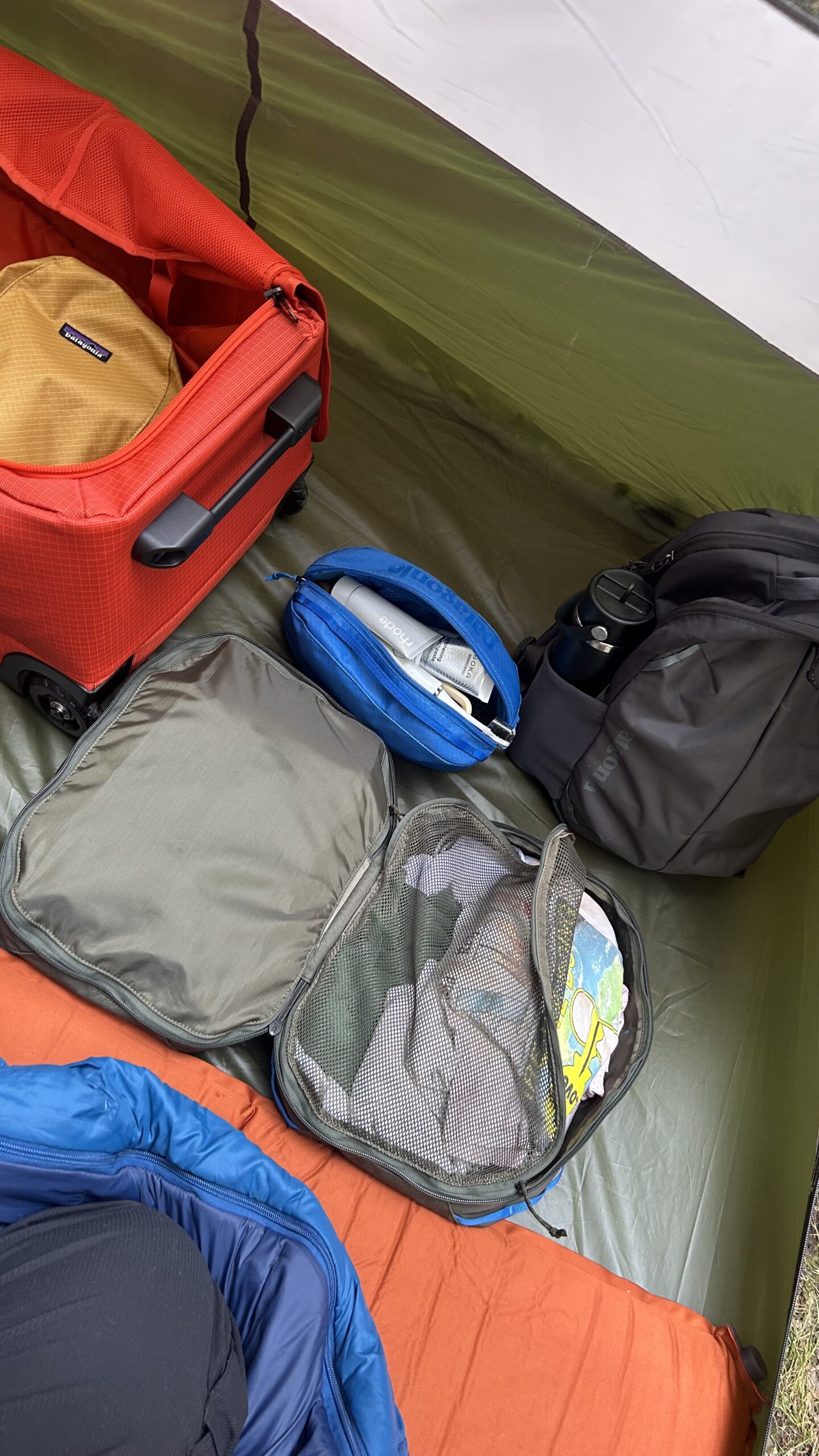 Inside a tent, a red suitcase, black bag, blue pouch, and a mesh packing cube with toiletries and a map rest on a green floor next to an orange sleeping bag.