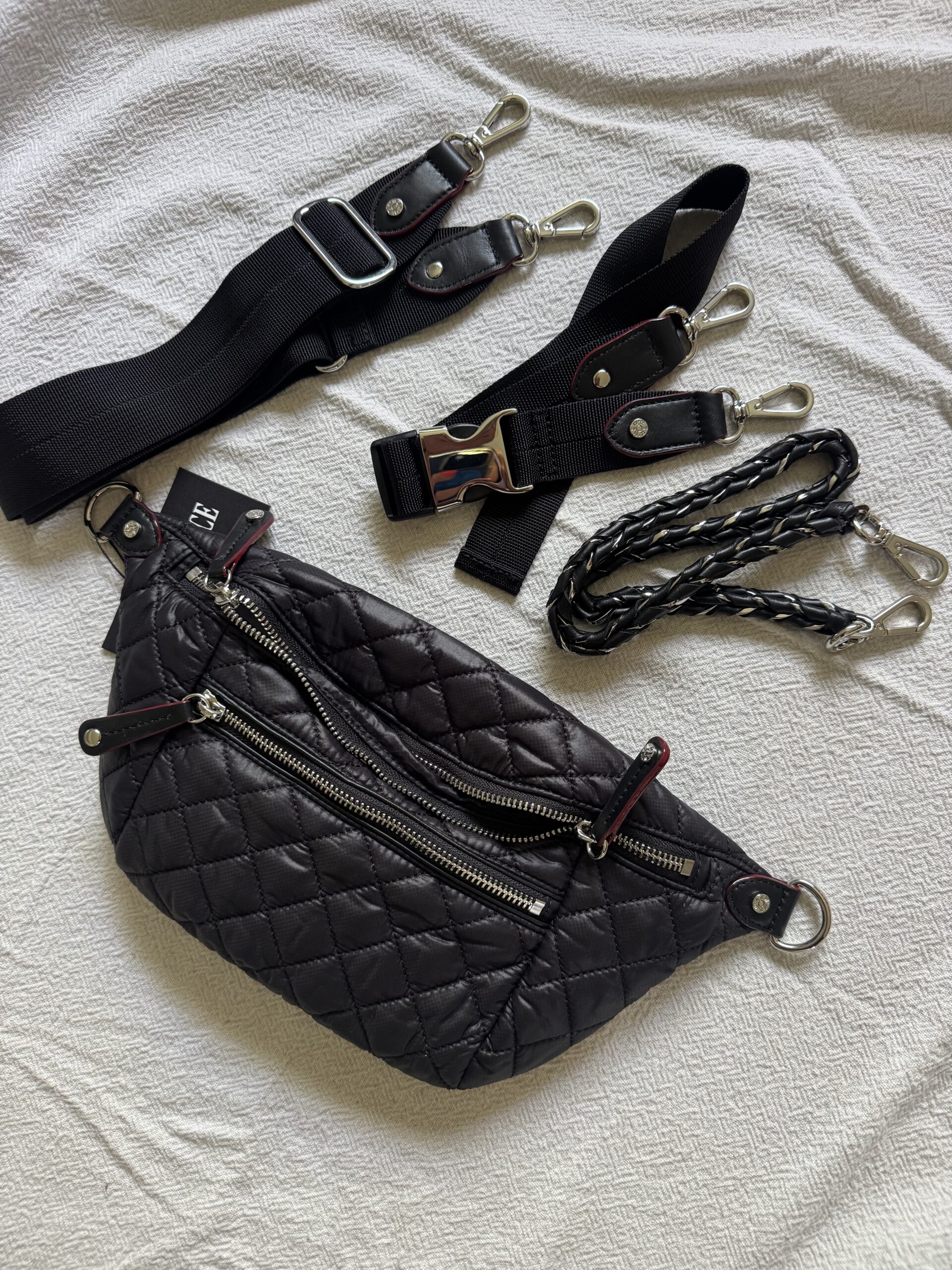 A black quilted bag with three detachable straps—one wide black strap, one seatbelt-style strap, and one braided strap—displayed on a textured white surface.