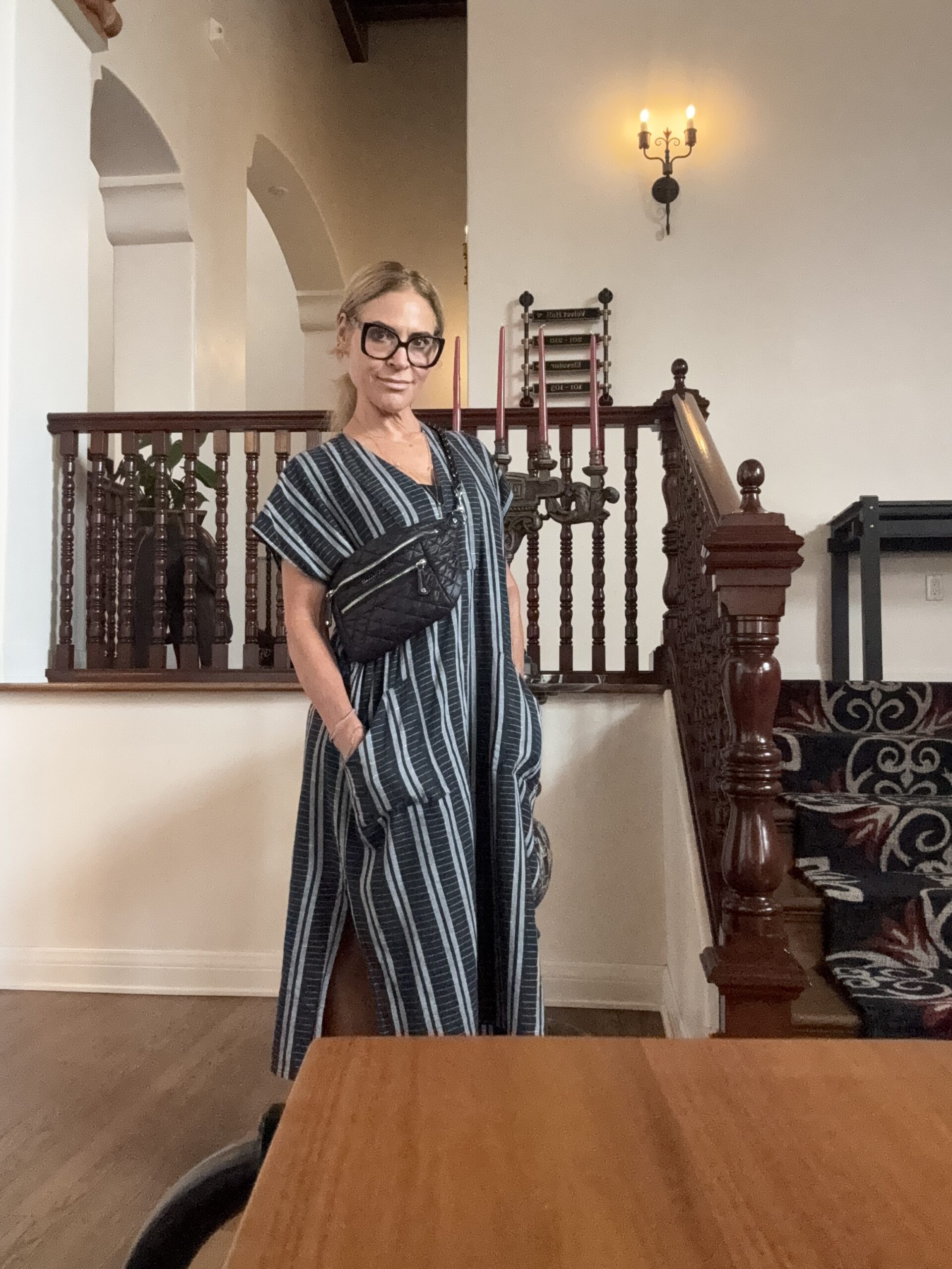 A woman in a striped dress stands indoors near a staircase, wearing glasses and a crossbody bag, with one hand in her pocket.