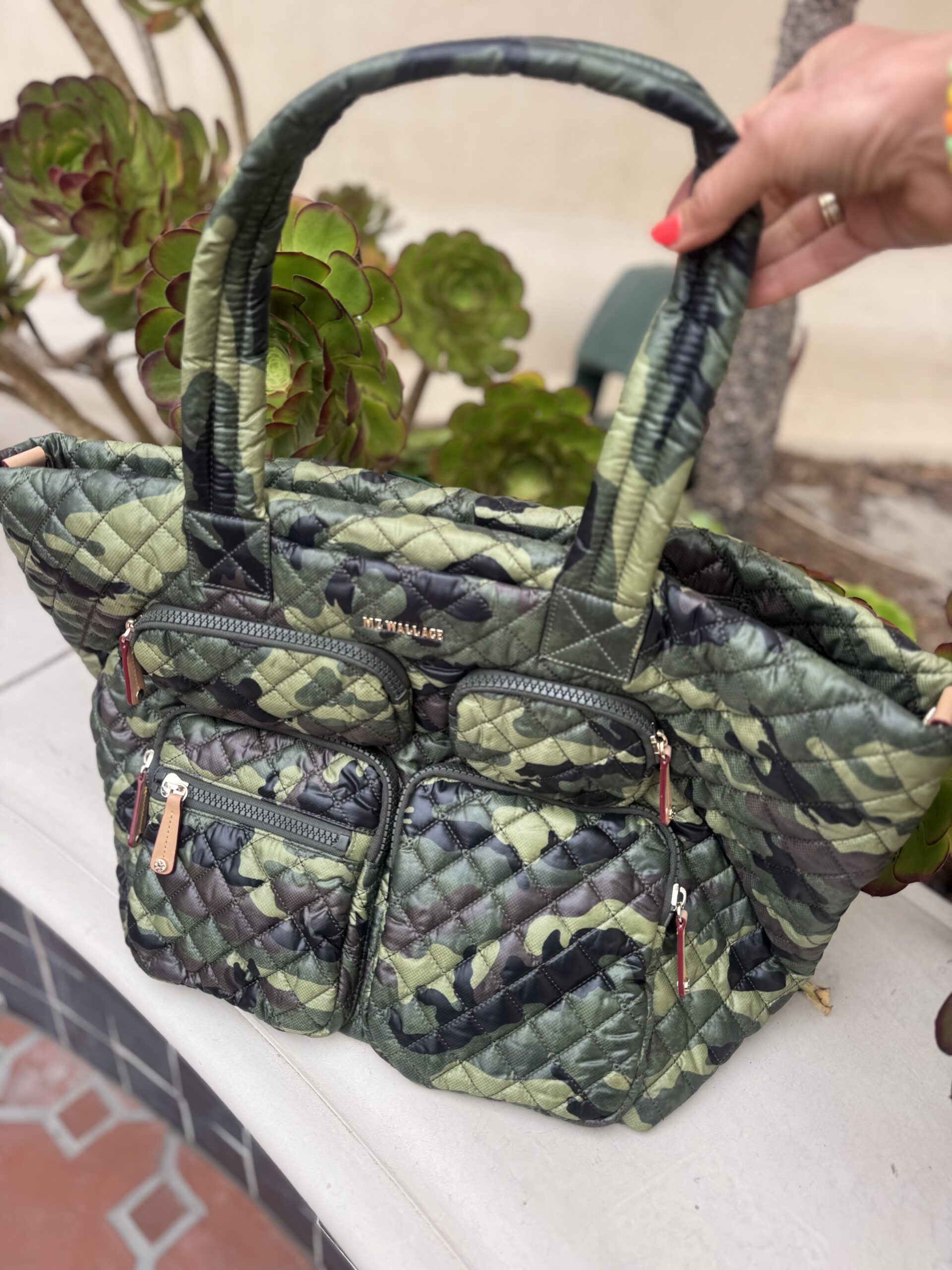A hand holds a quilted green camouflage tote bag with multiple exterior zippered pockets, against an outdoor background with plants and a tiled surface.