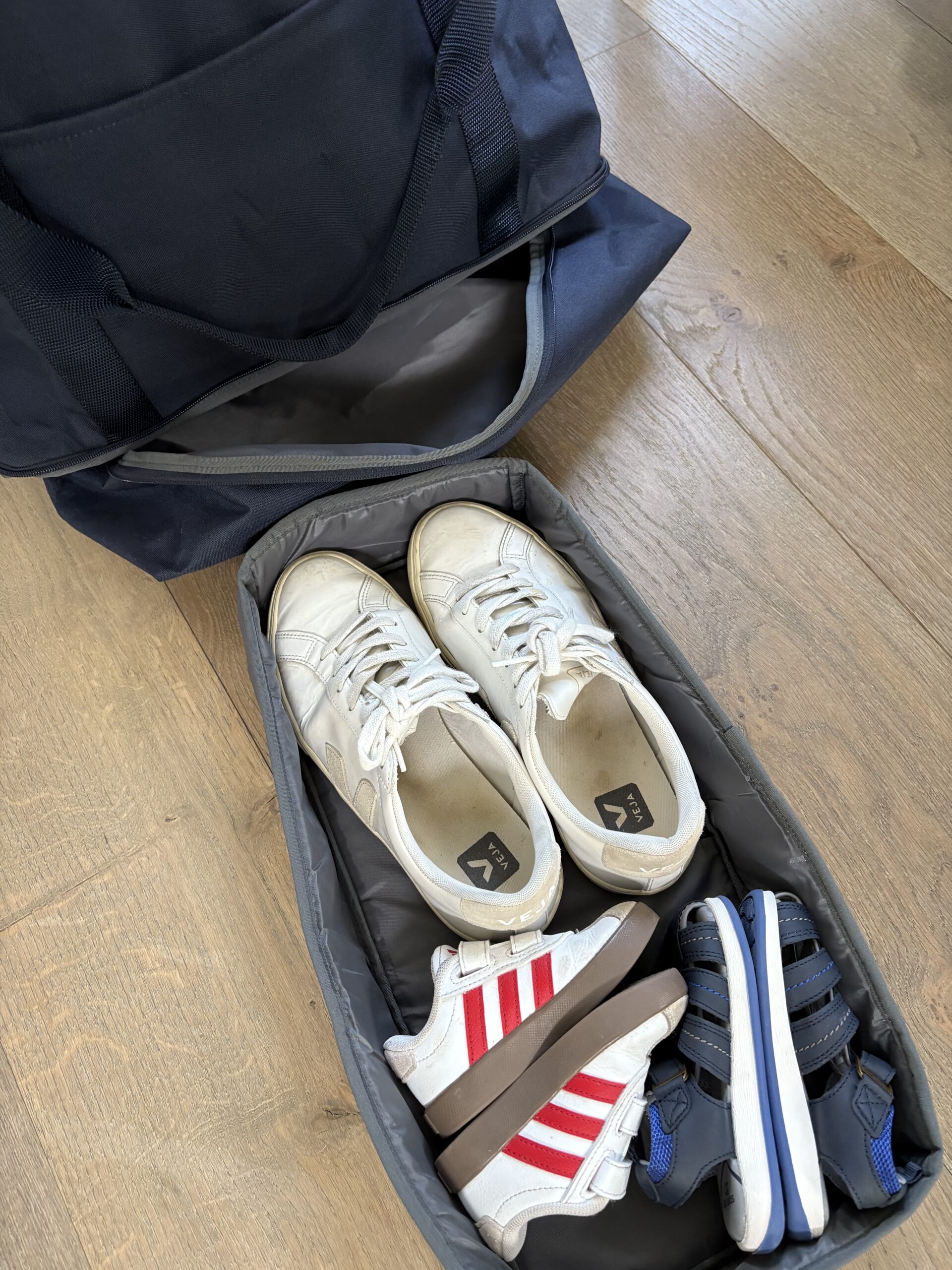 A gym bag sits open on a wooden floor, revealing a compartment holding a pair of adult white sneakers, a pair of kids' sandals, and a pair of kids' sneakers with red stripes.