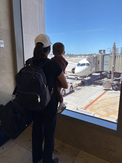 An adult holding a child looks out an airport window at a passenger airplane parked at a gate on a sunny day.