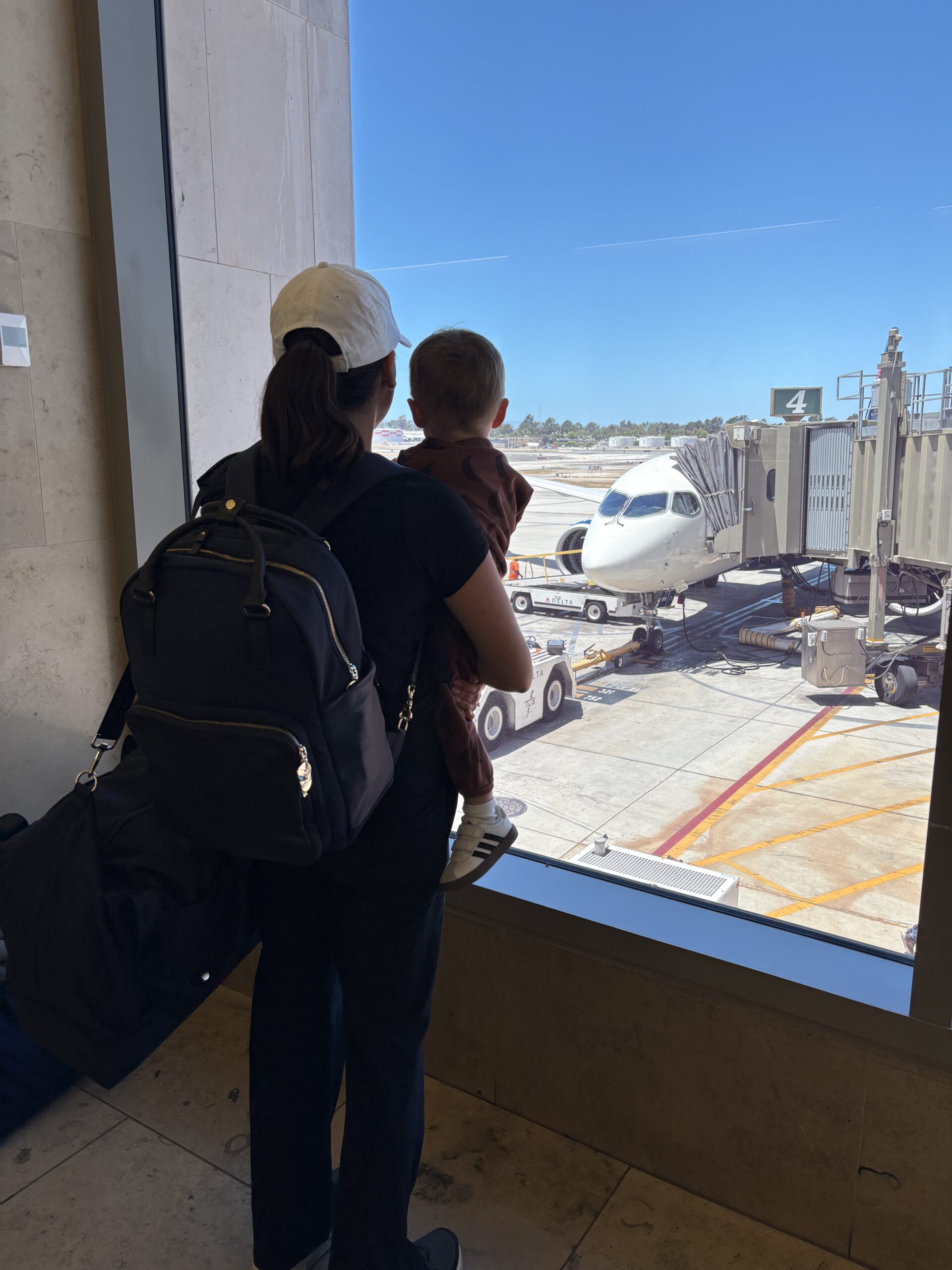 An adult holding a child looks out an airport window at a passenger airplane parked at a gate on a sunny day.