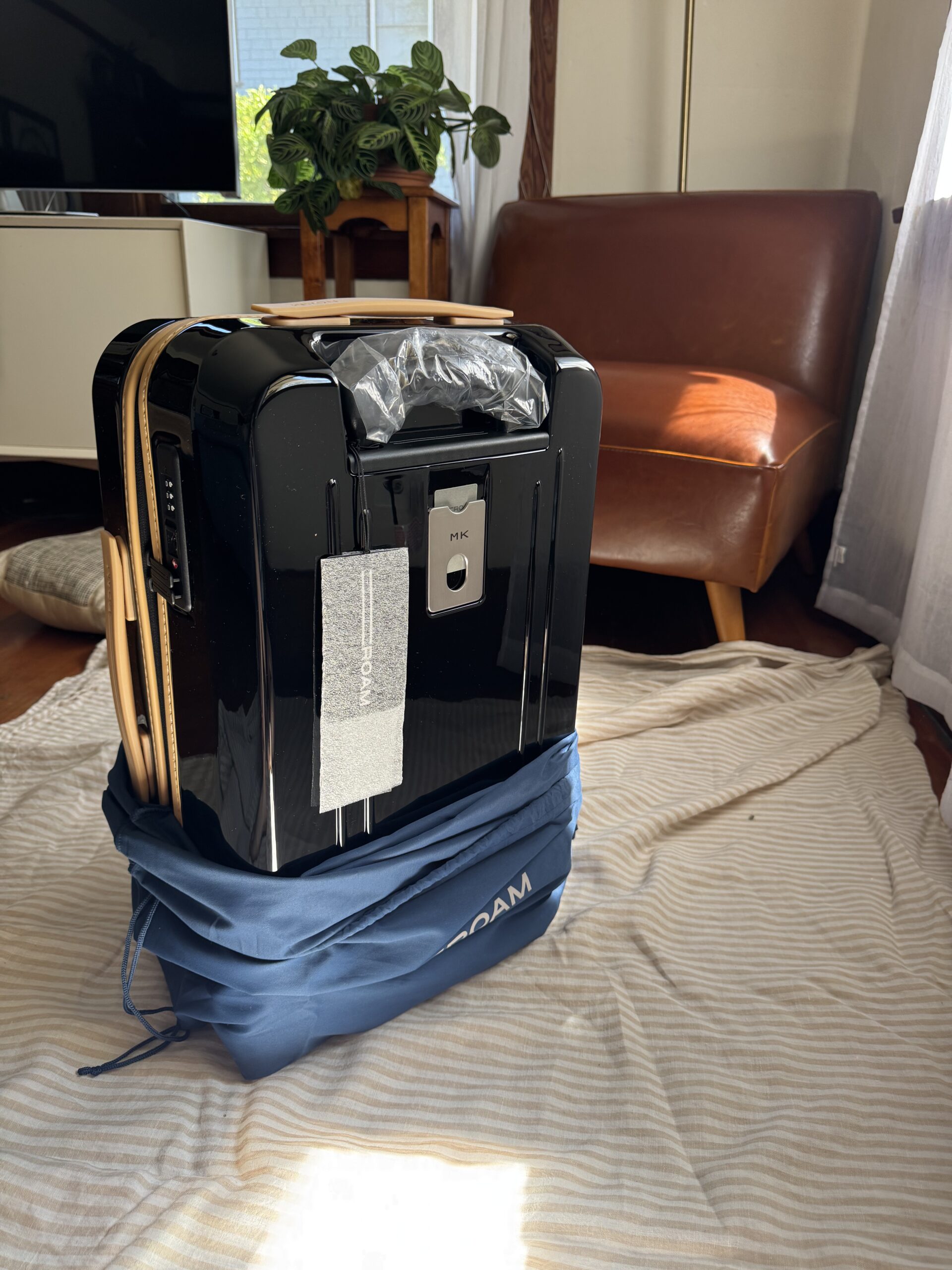 A black hard-shell suitcase stands upright on a striped cloth, partially wrapped in a blue bag, with a chair and houseplant in the background.
