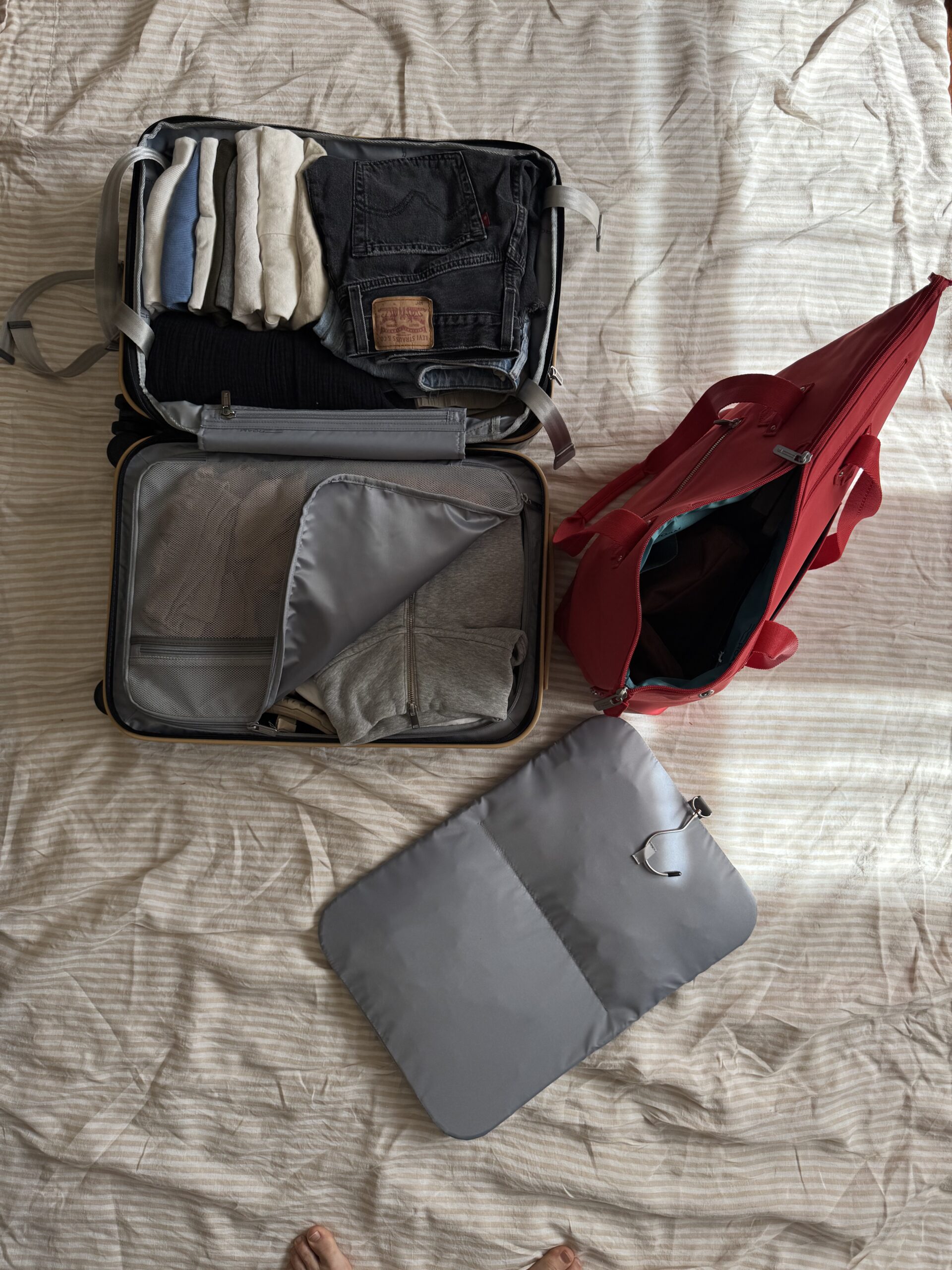 An open suitcase with neatly folded clothes, a red bag, and a gray packing pouch are laid out on a beige striped bedspread, with feet visible at the bottom of the image.
