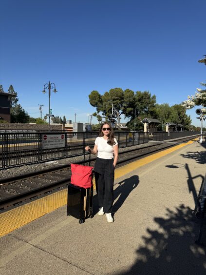 A woman stands on a train station platform with a suitcase and red bag, wearing sunglasses, a white top, and black pants. The sky is clear and sunny.