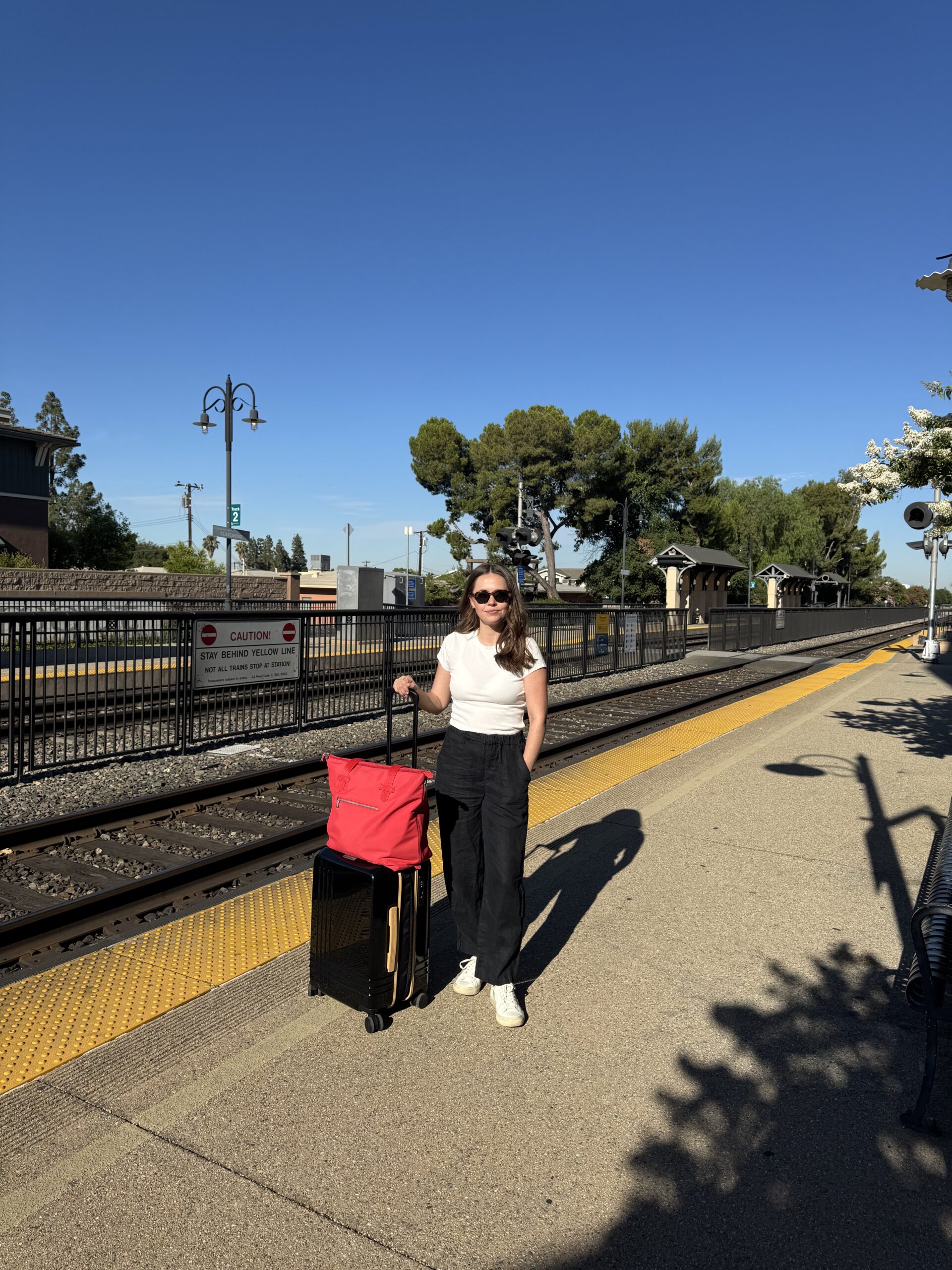 A woman stands on a train station platform with a suitcase and red bag, wearing sunglasses, a white top, and black pants. The sky is clear and sunny.