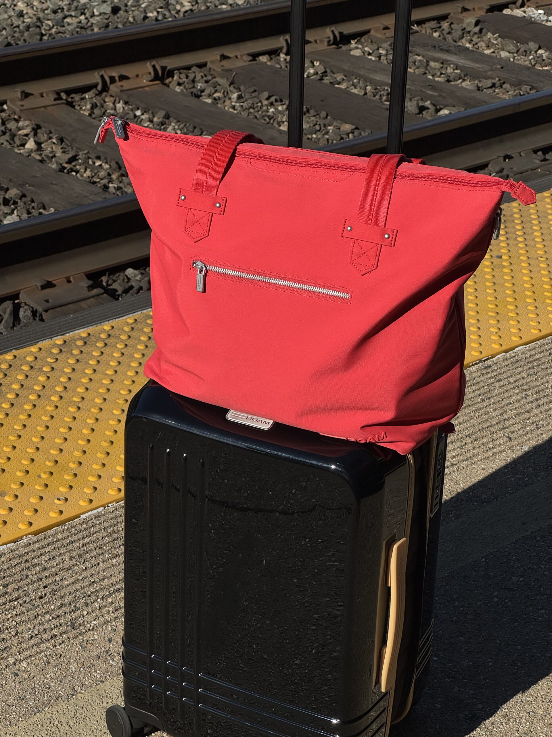 A red tote bag sits on top of a black rolling suitcase on a train station platform near railway tracks.